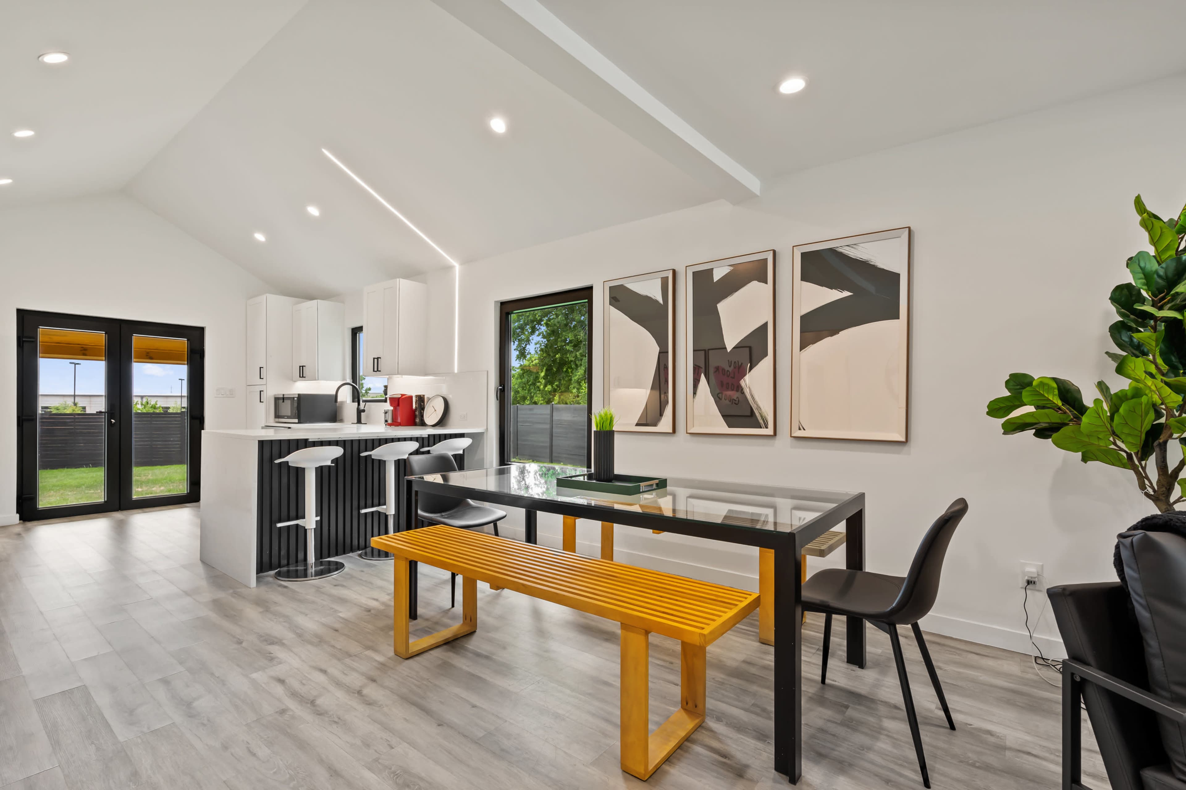 A modern kitchen and dining area featuring a glass dining table with a yellow bench, white cabinetry, and a black countertop.