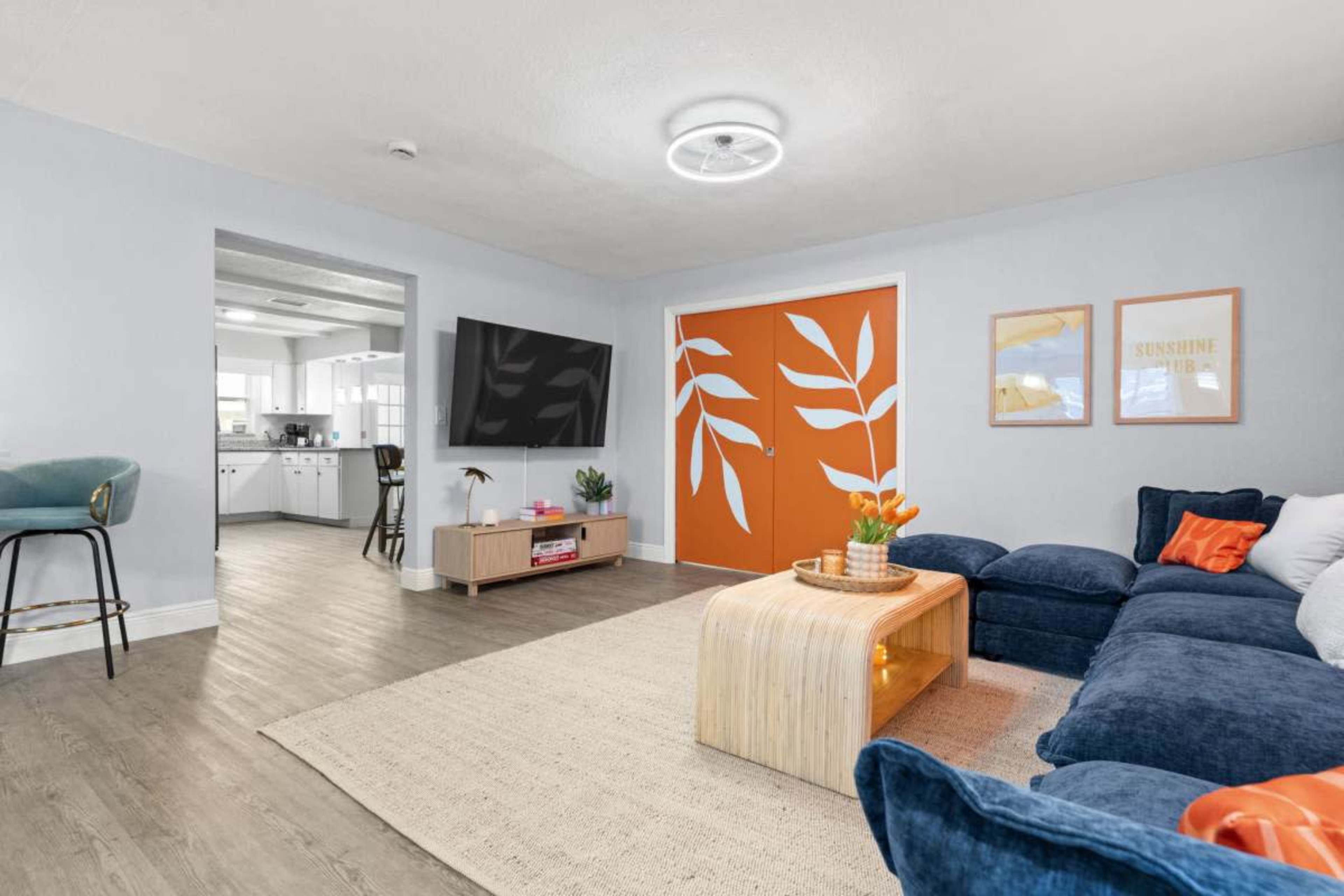 The image shows a living room with a dark blue sectional sofa, a wooden coffee table, an orange accent wall with leaf designs, and a television mounted on the opposite wall.