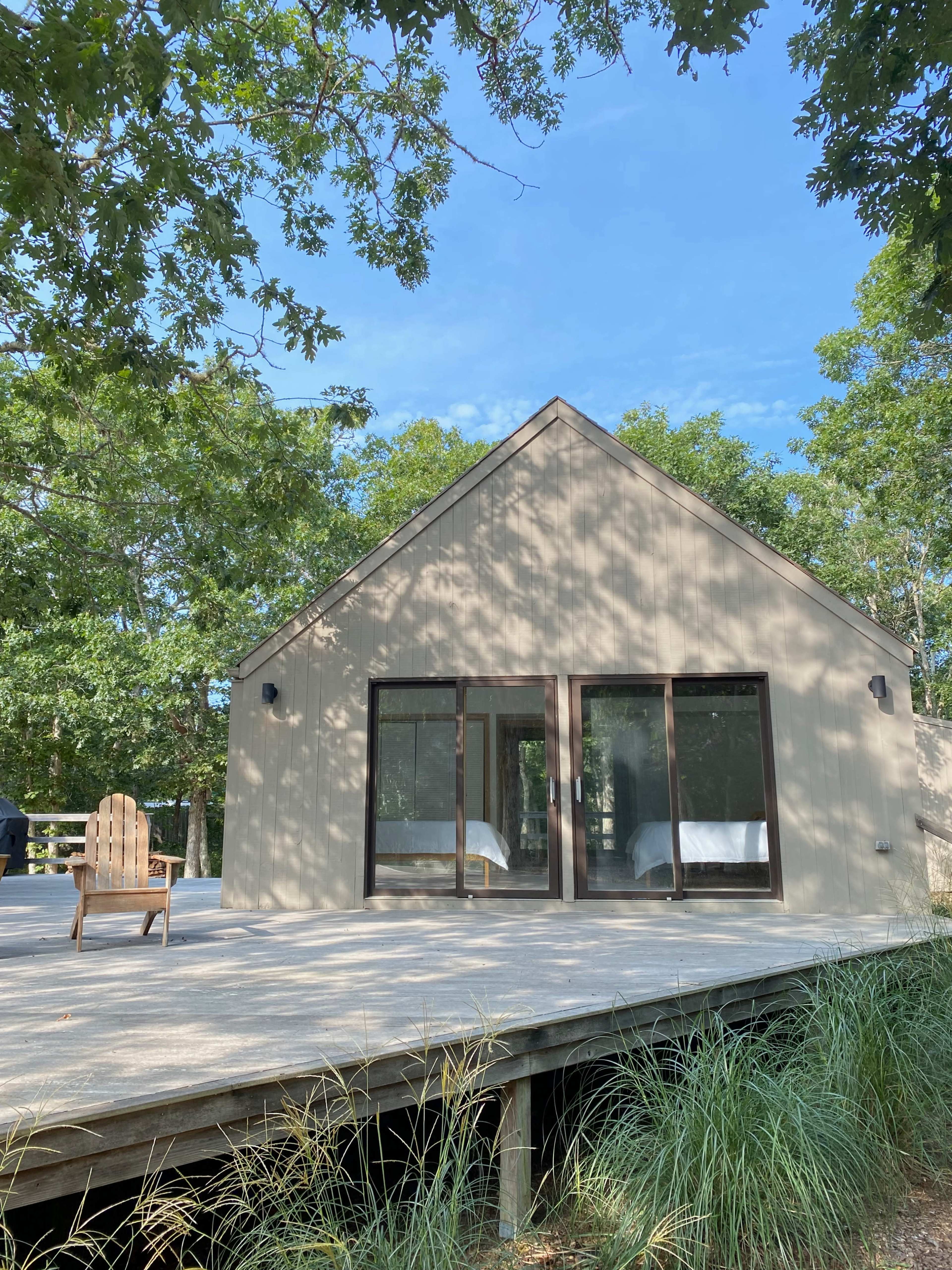 A modern, minimalist house with large windows is surrounded by trees and features a wooden deck with an Adirondack chair in the foreground.