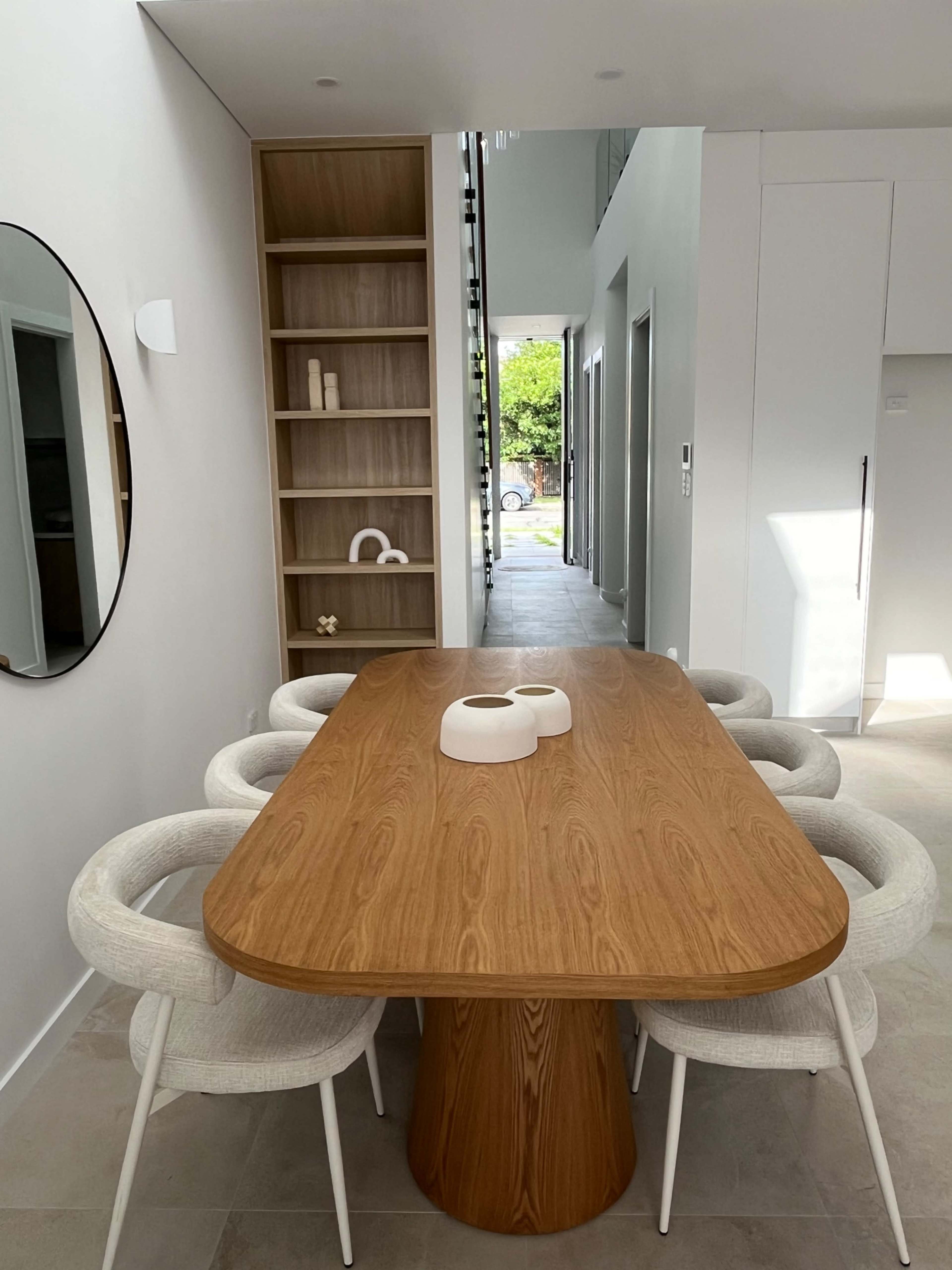 A wooden dining table with six white chairs is positioned in a modern, well-lit dining area, adjacent to a shelf displaying decorative items.
