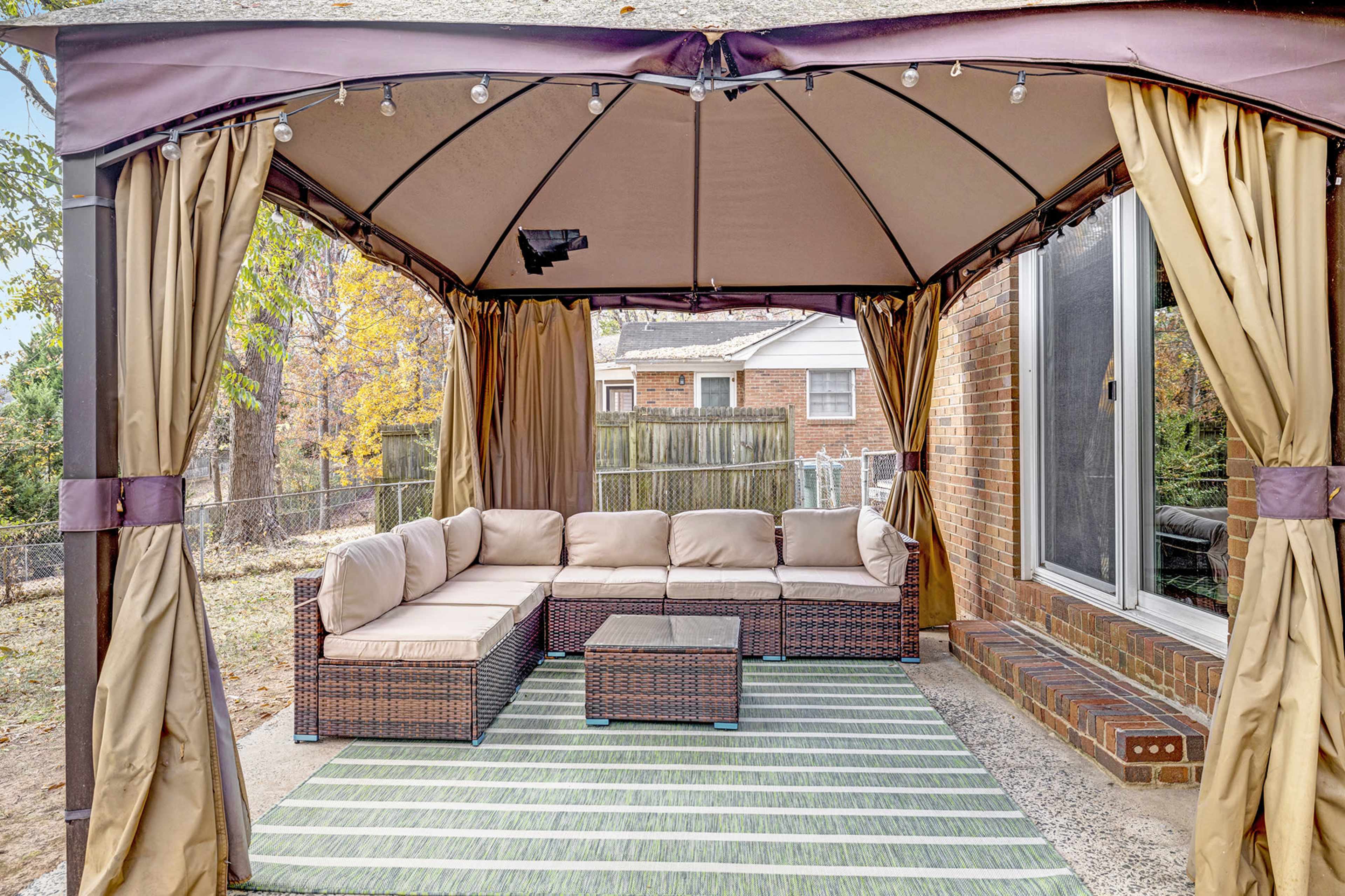 A covered patio area features a sectional sofa and a coffee table on a striped rug, with a backdrop of a fenced yard and neighboring houses.