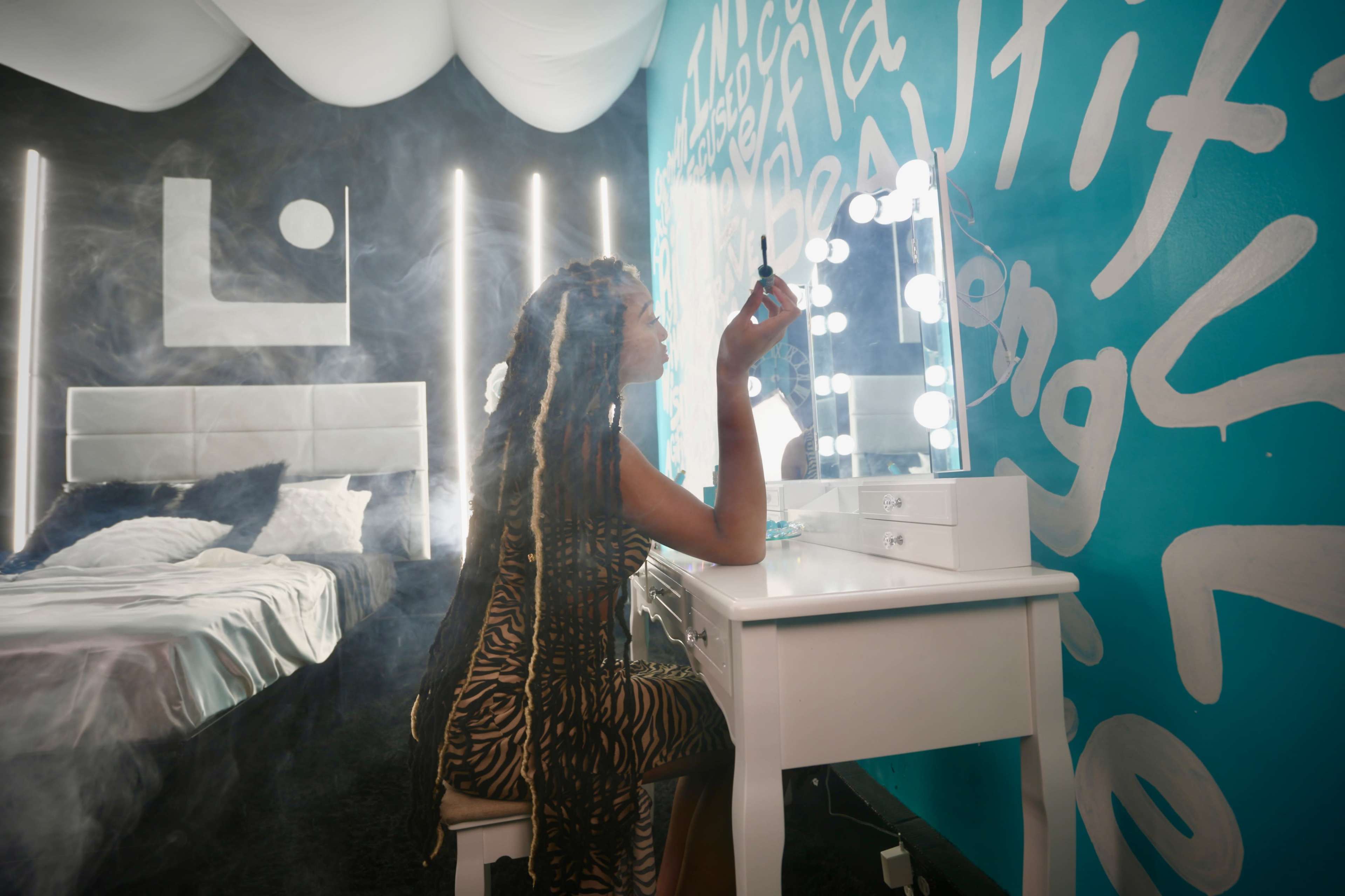 A woman with long braided hair sits at a white vanity in a room with a brightly colored wall and soft bed linens.