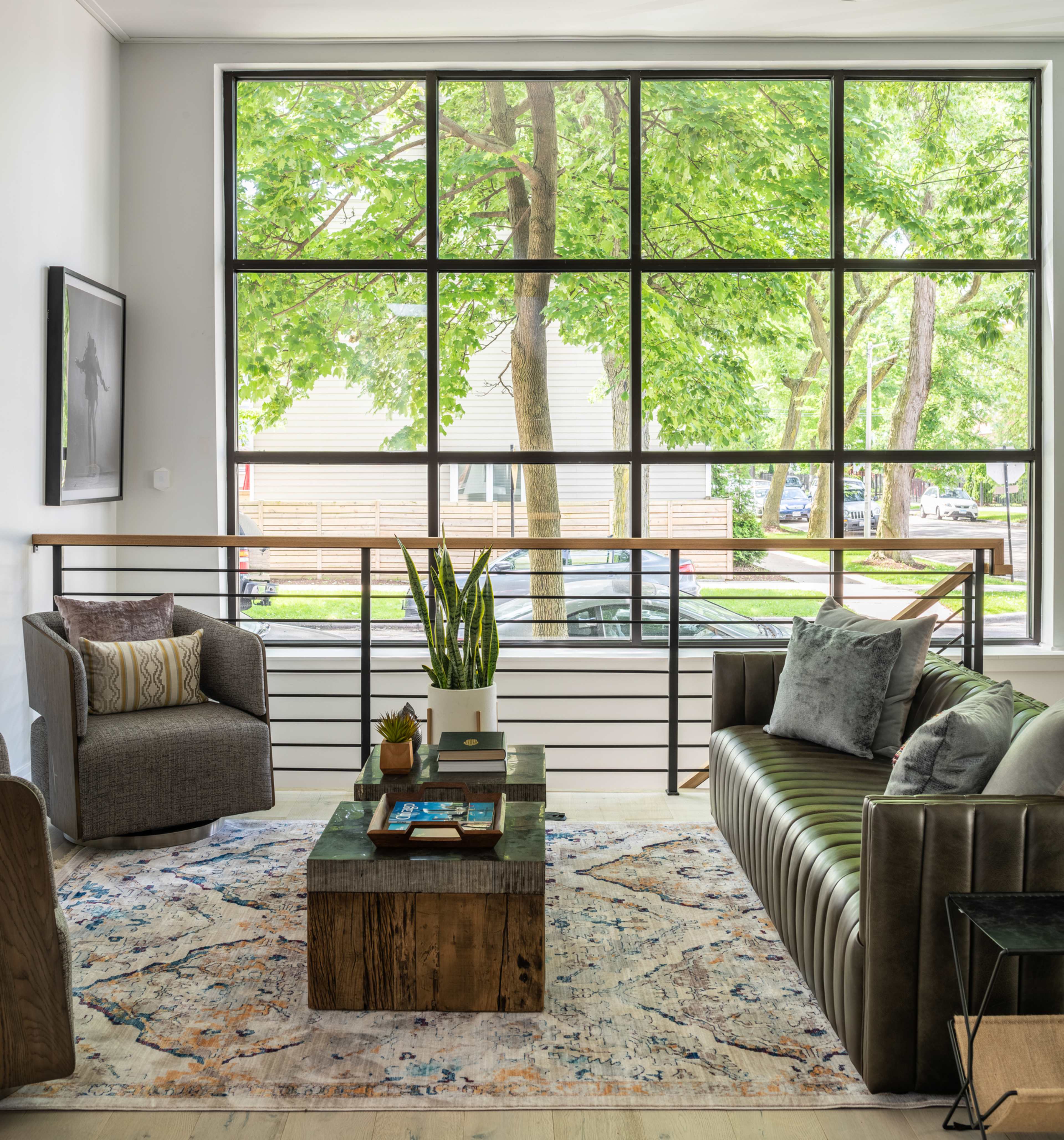 A modern living room features a green leather sofa, gray armchairs, a wooden coffee table, and large windows overlooking trees outside.