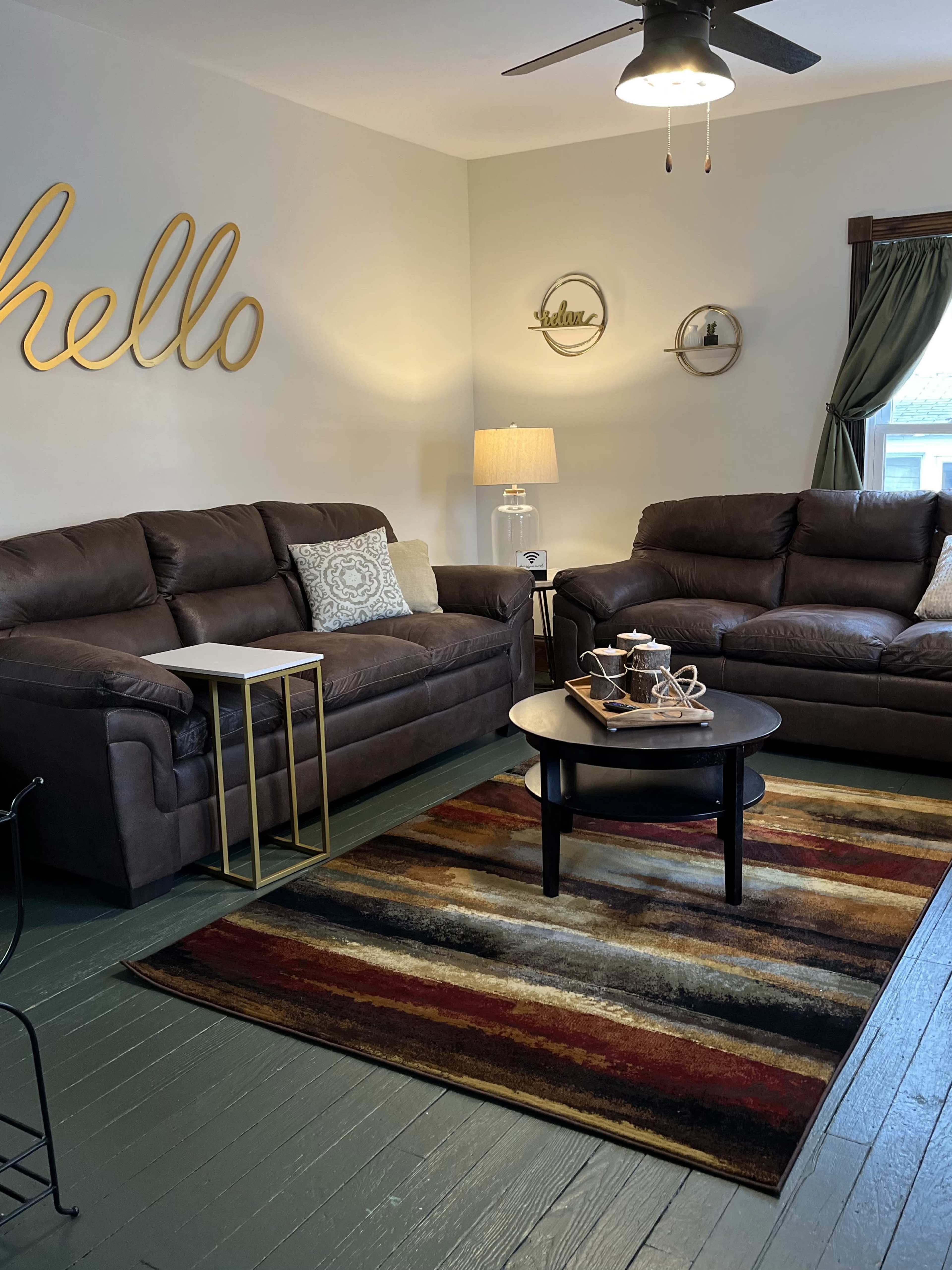 A living room features two brown sofas, a round black coffee table, a striped area rug, and decor on the walls.