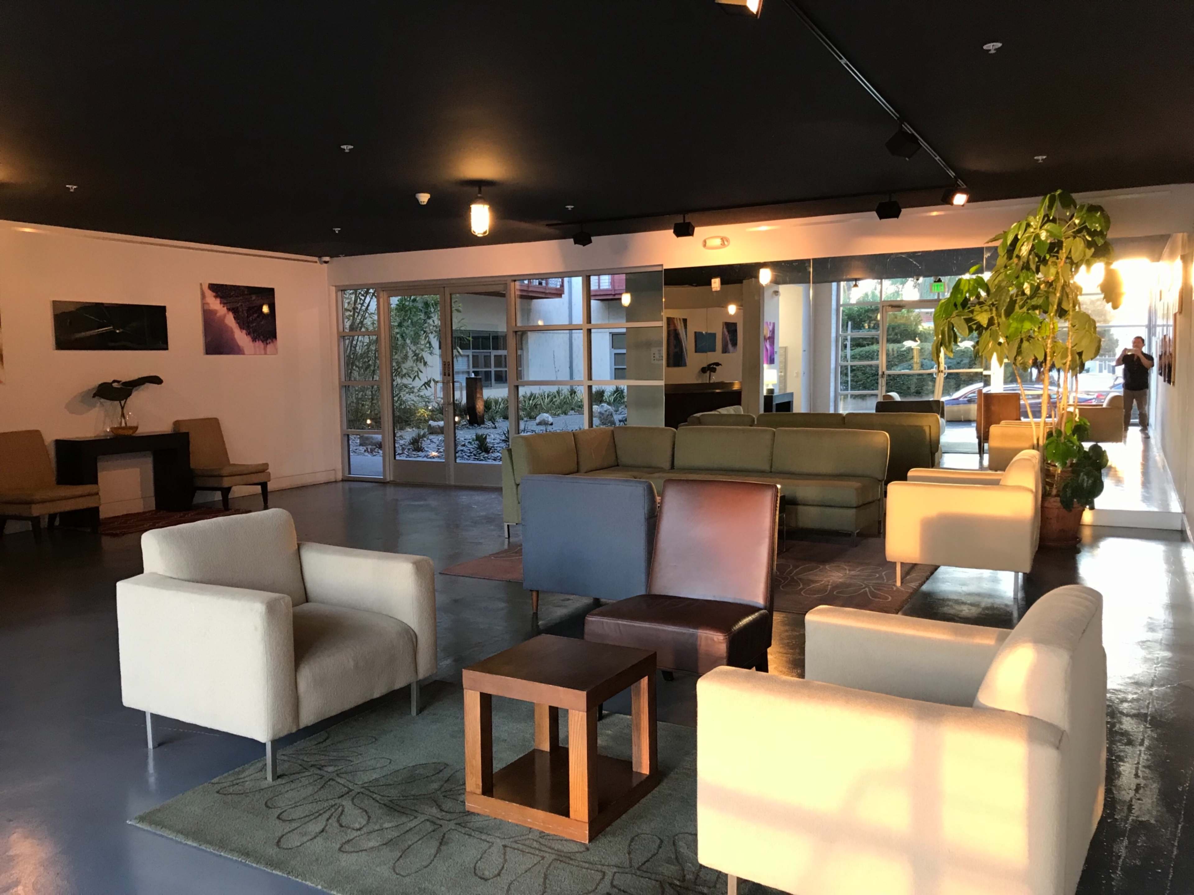 The image shows a modern lounge area with various seating arrangements, including chairs and sofas, and large windows that allow natural light to enter.