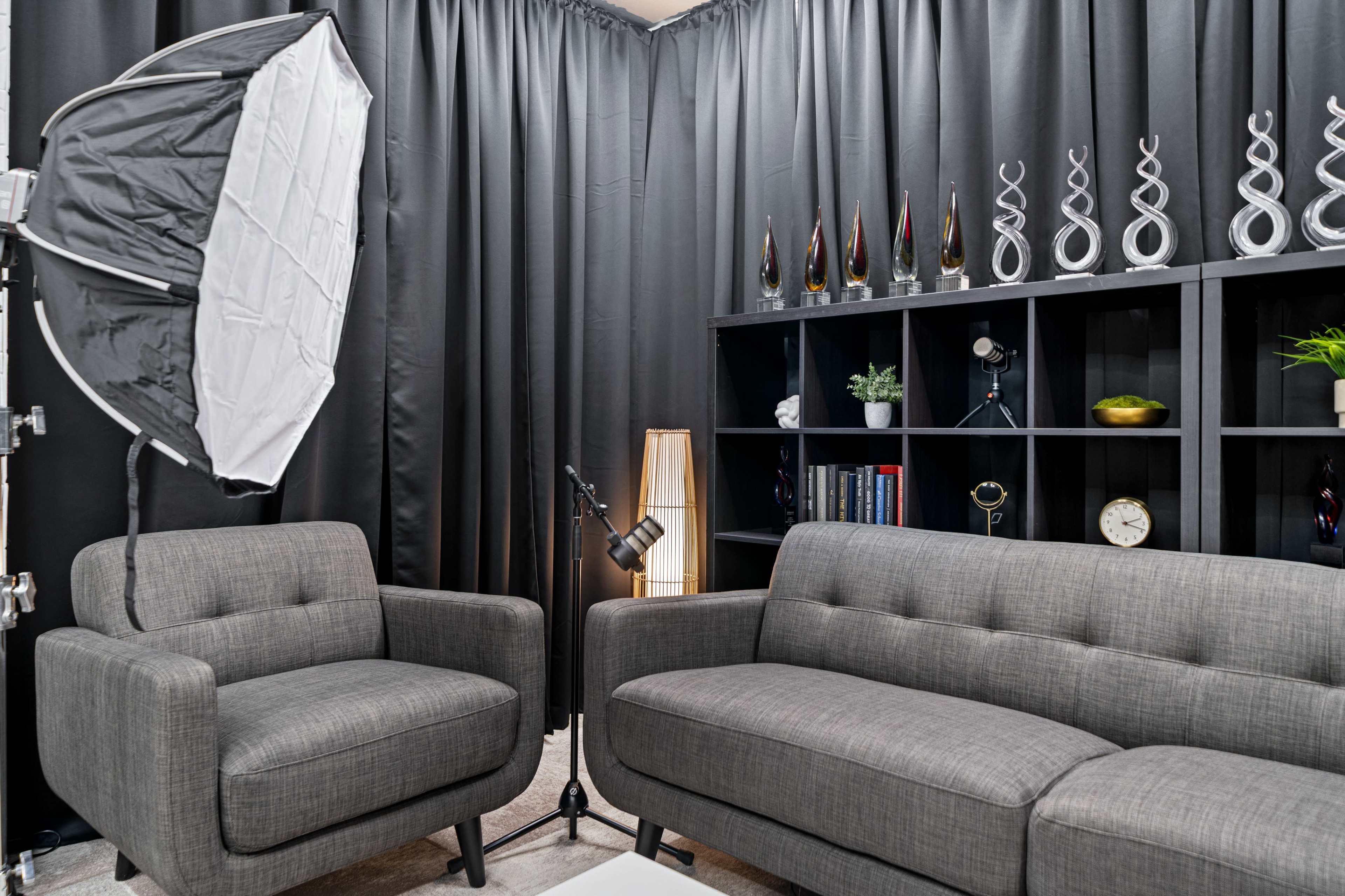 The image shows a modern sitting area featuring two gray sofas, a black bookshelf with decorative items, and soft lighting against a backdrop of black curtains.