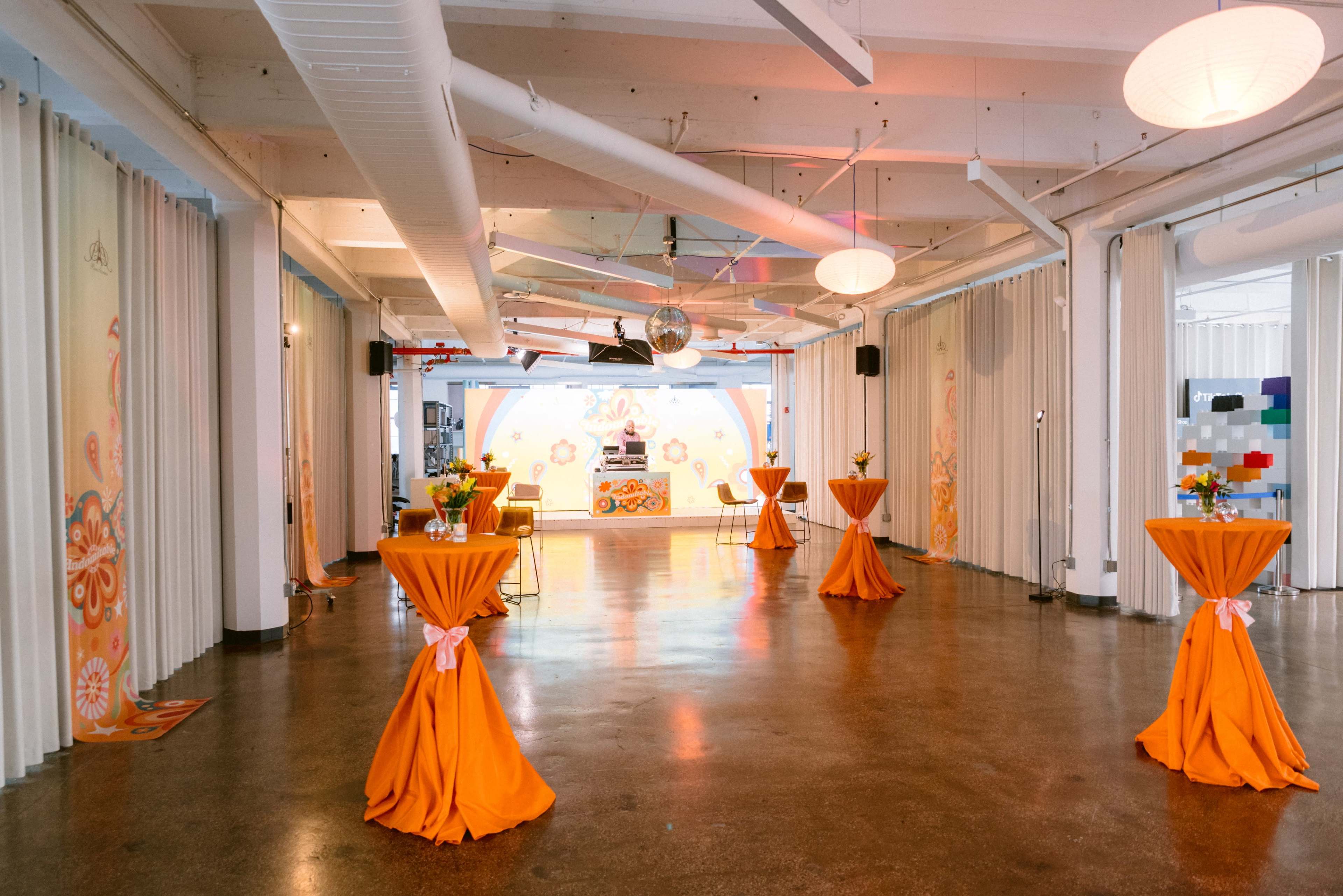 The spacious event venue features orange draped tables and colorful decorative elements against a bright backdrop.