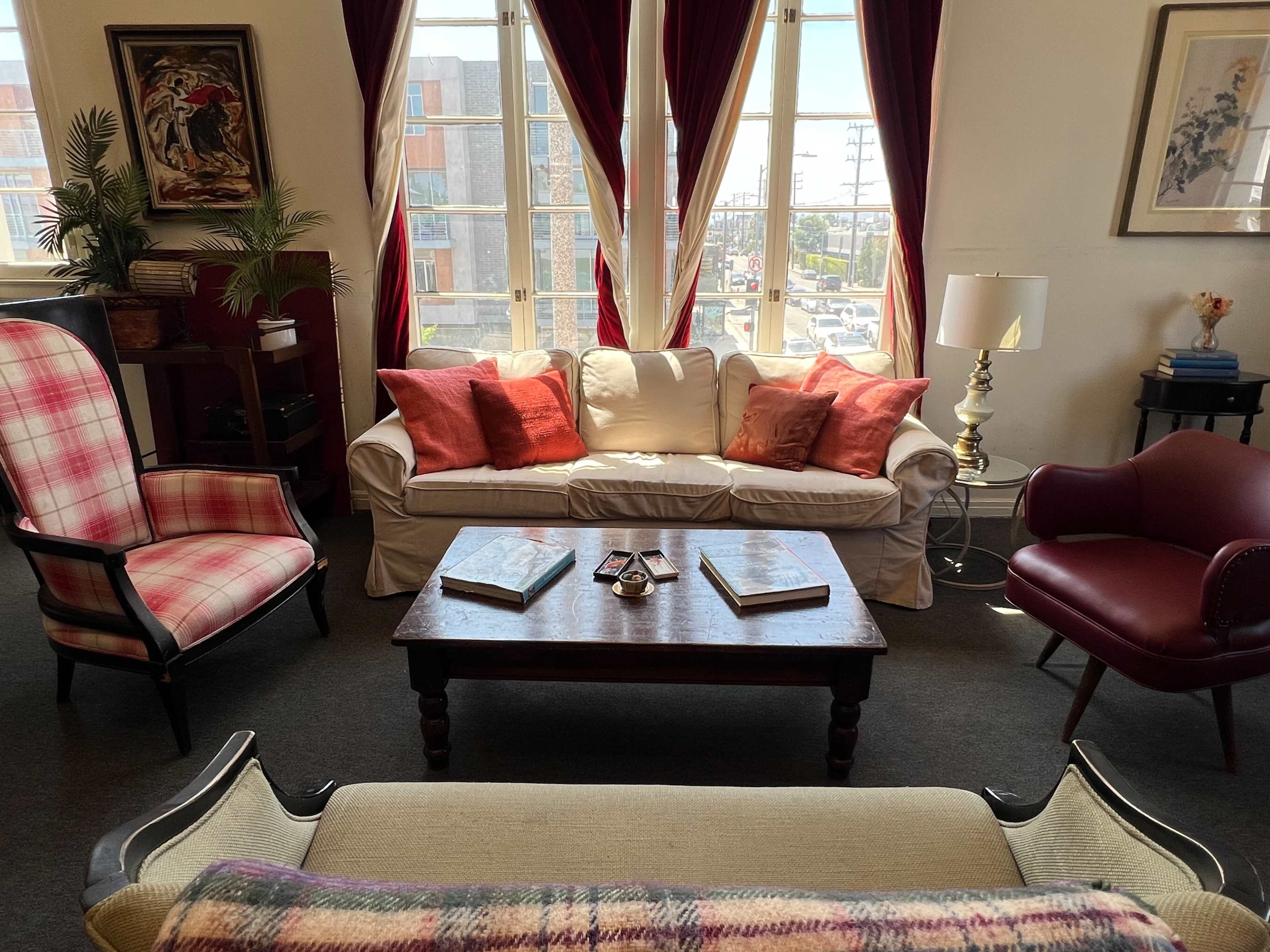 A cozy living room with a white sofa, two armchairs, a wooden coffee table, and bright windows dressed with red curtains.