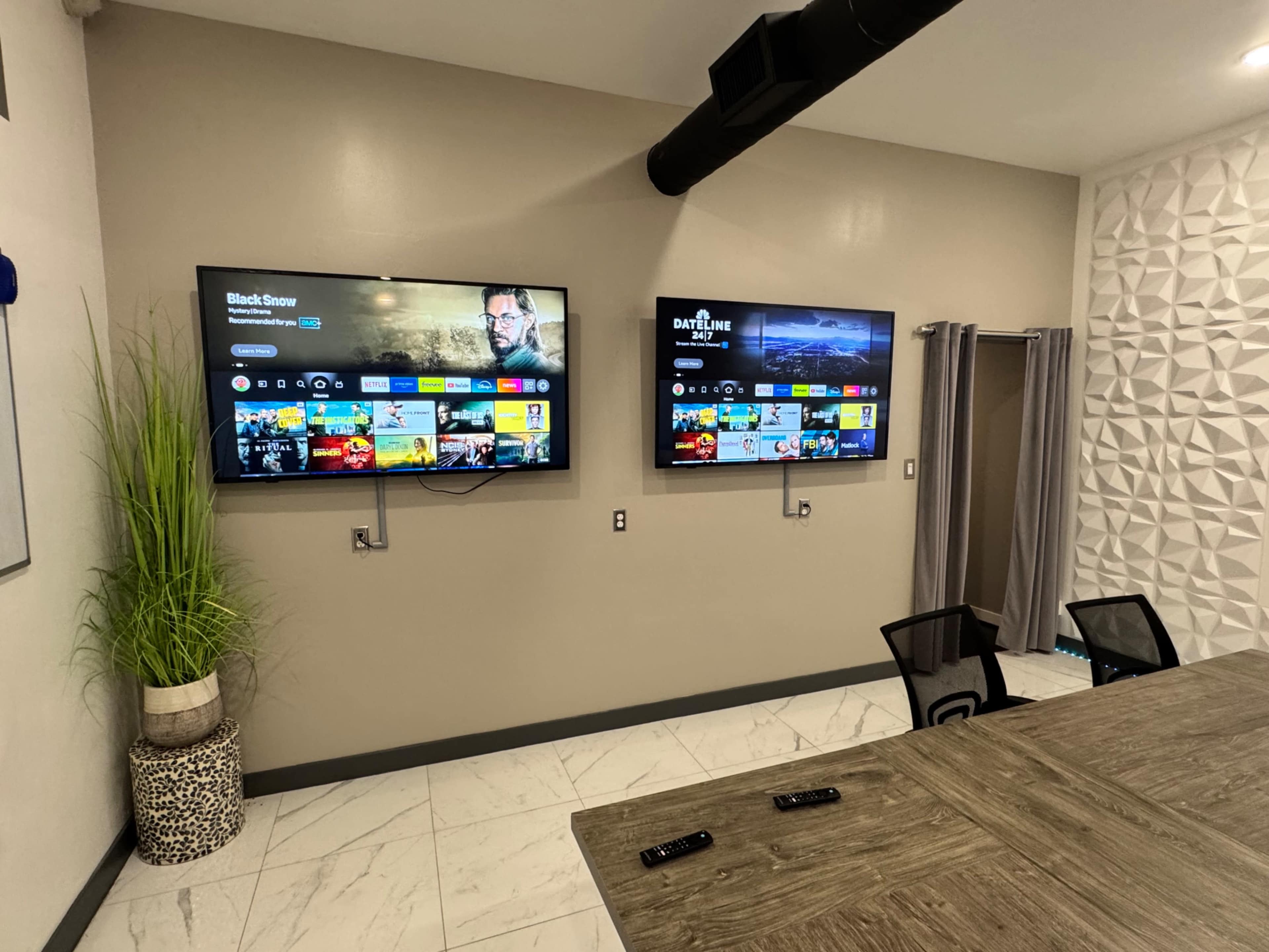 The image shows a modern room with two wall-mounted flat-screen TVs displaying streaming apps, alongside a table and chairs, and decorative plants in a planter.