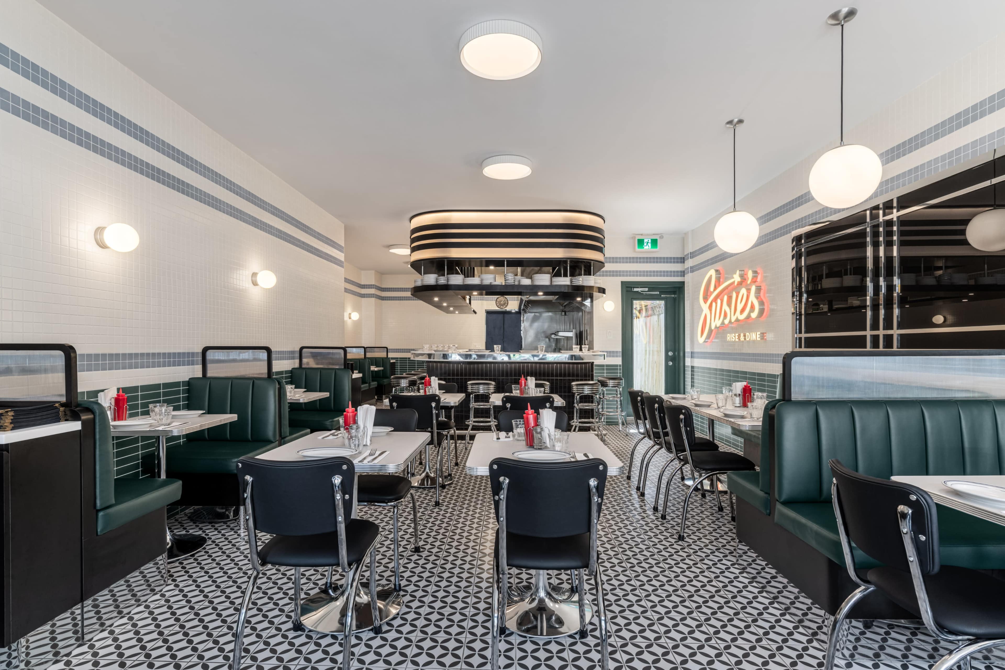The image shows a retro-style diner with black and green seating, white tile walls, and a patterned floor, featuring a counter and an open kitchen area.