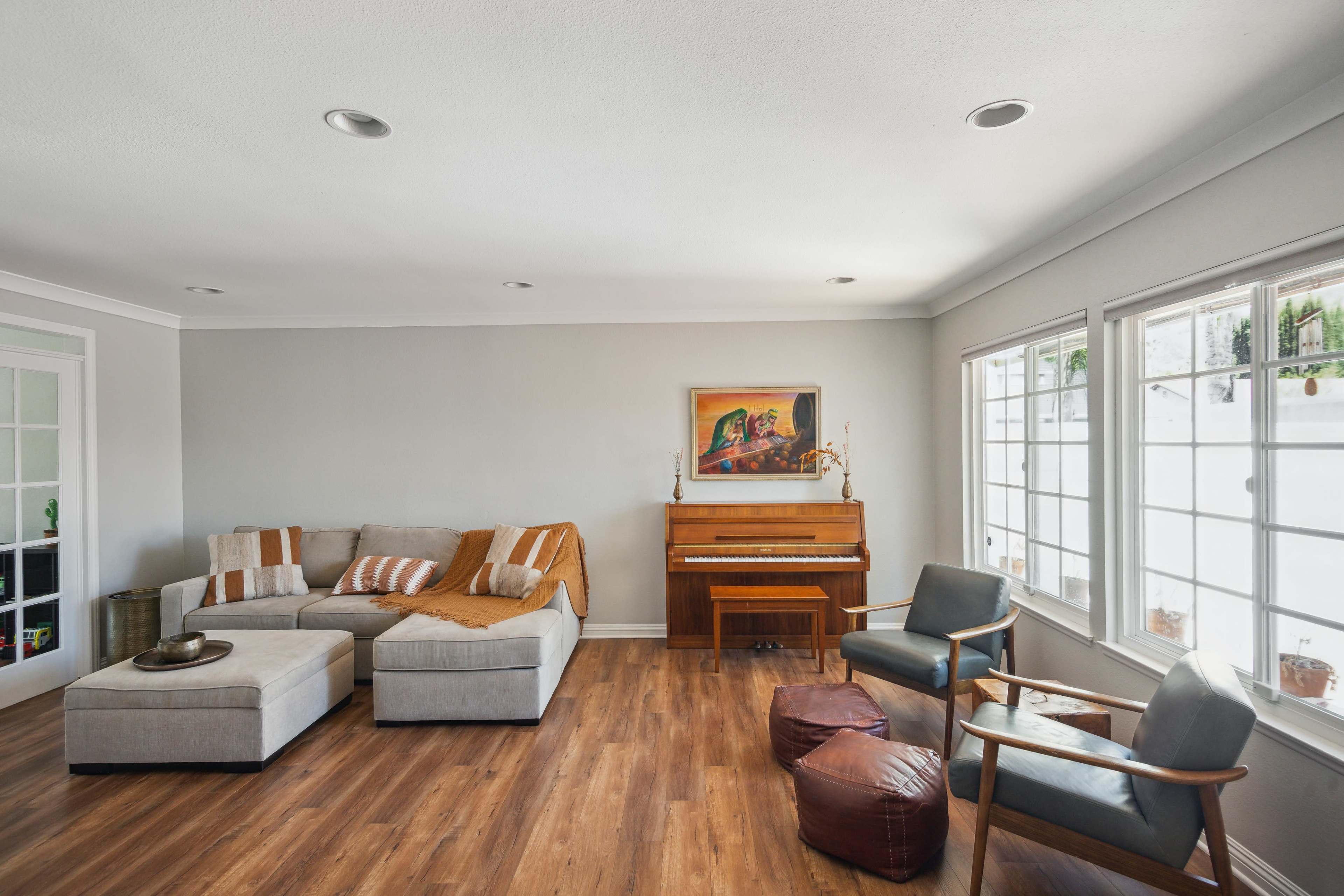 A light-filled living room features a sectional sofa, a wooden piano, and chairs arranged around a coffee table.