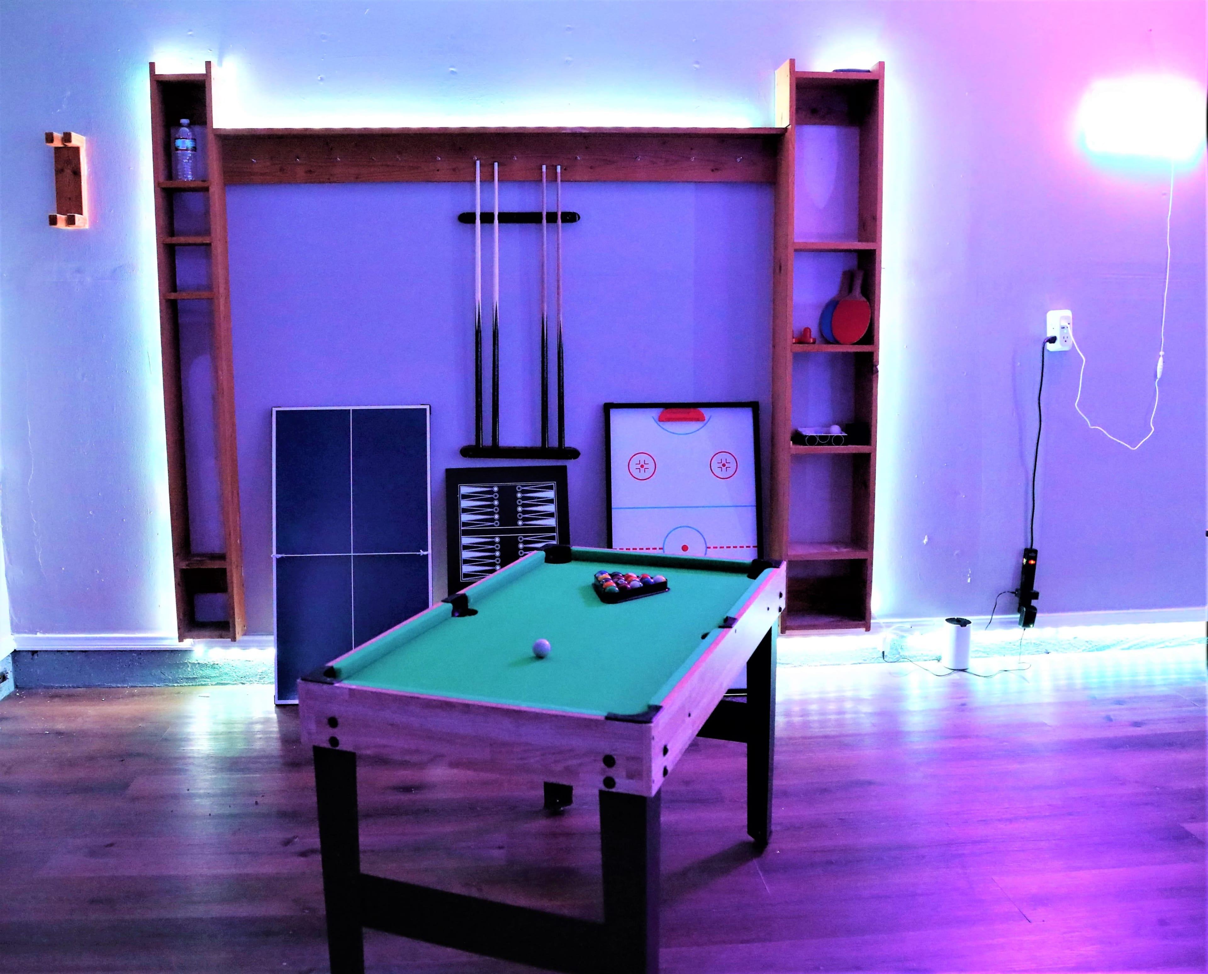 A game room setup featuring a pool table, a small air hockey table, and shelves against a wall with ambient lighting.