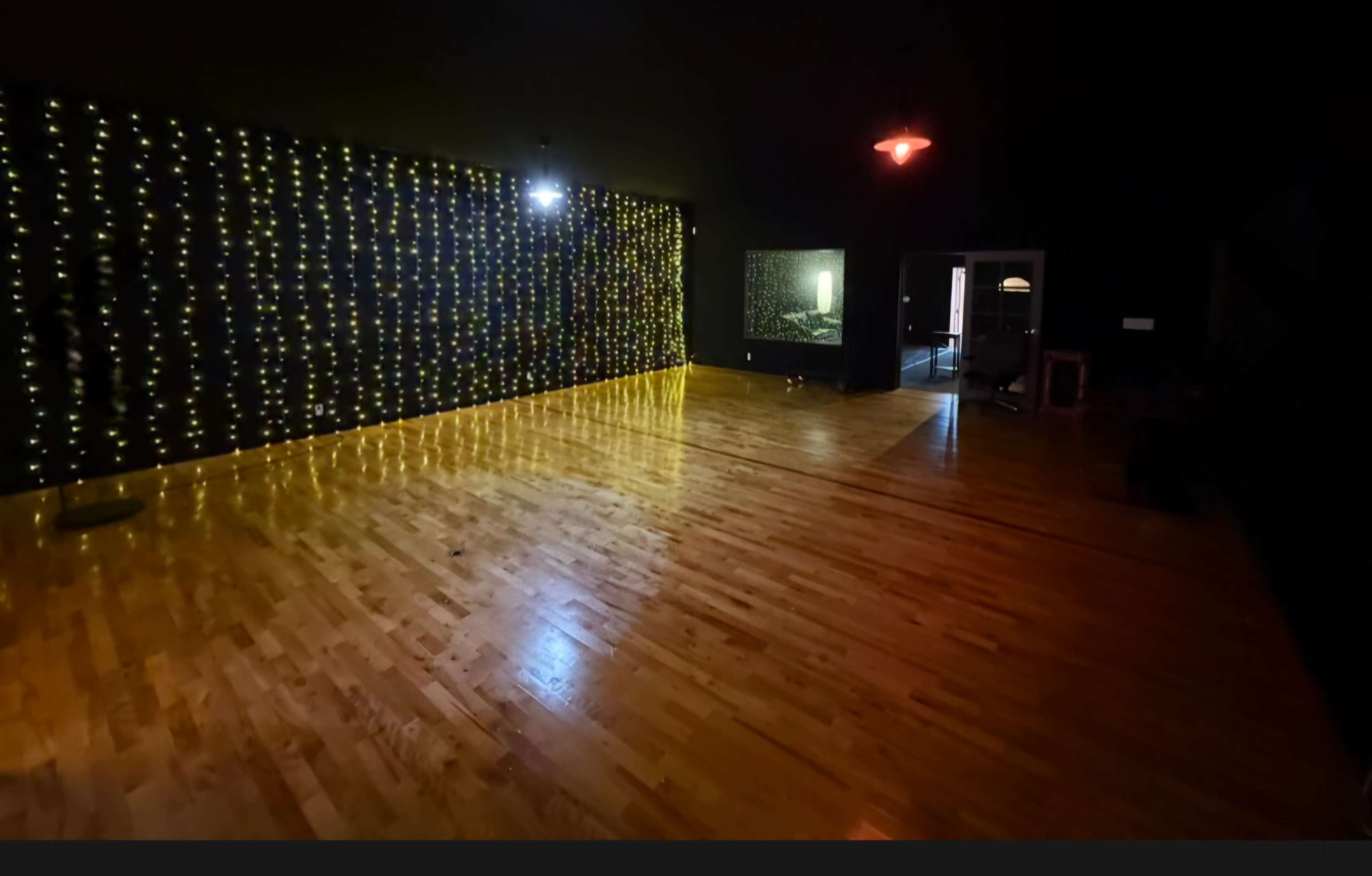 The image shows a sparsely furnished dance studio with polished wooden floors and a wall adorned with string lights.