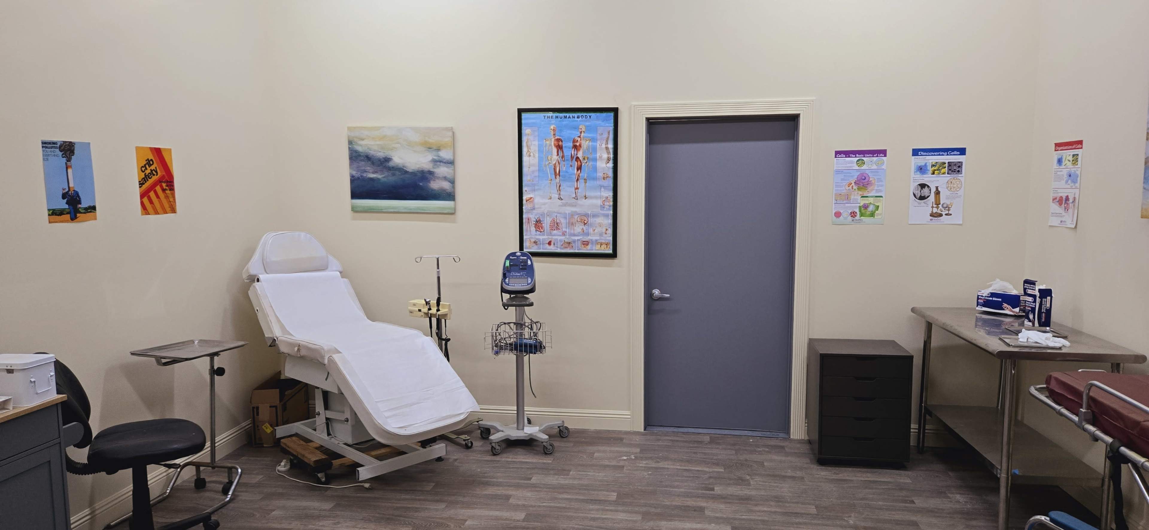 A clinical examination room featuring an adjustable examination table, a rolling cart, a desk, and various posters on the walls.