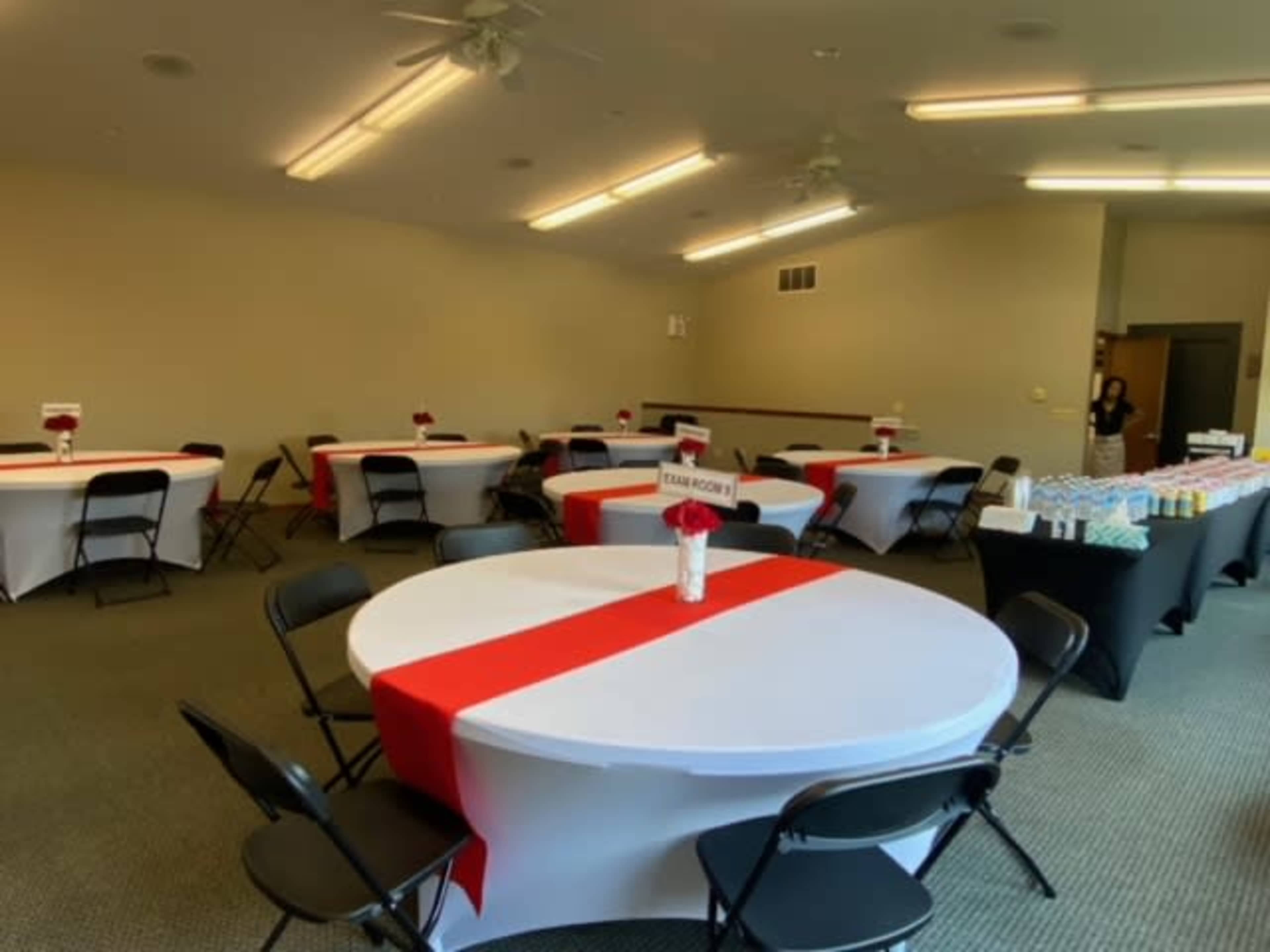 A banquet room with several round and rectangular tables set with red and white table covers, arranged for an event.
