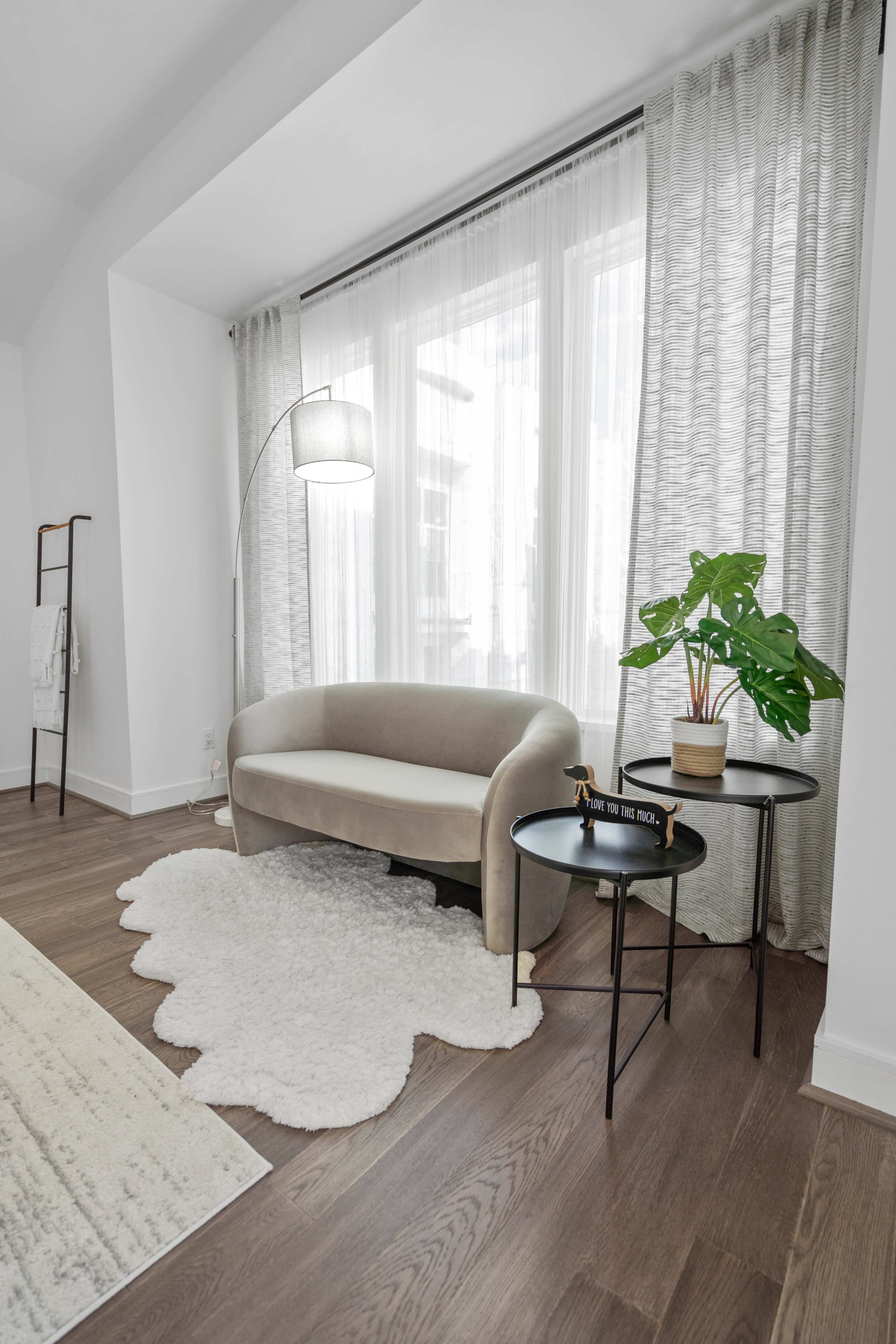A soft-textured, light-colored sofa is positioned next to a small black table and a potted plant in a well-lit room with sheer curtains.