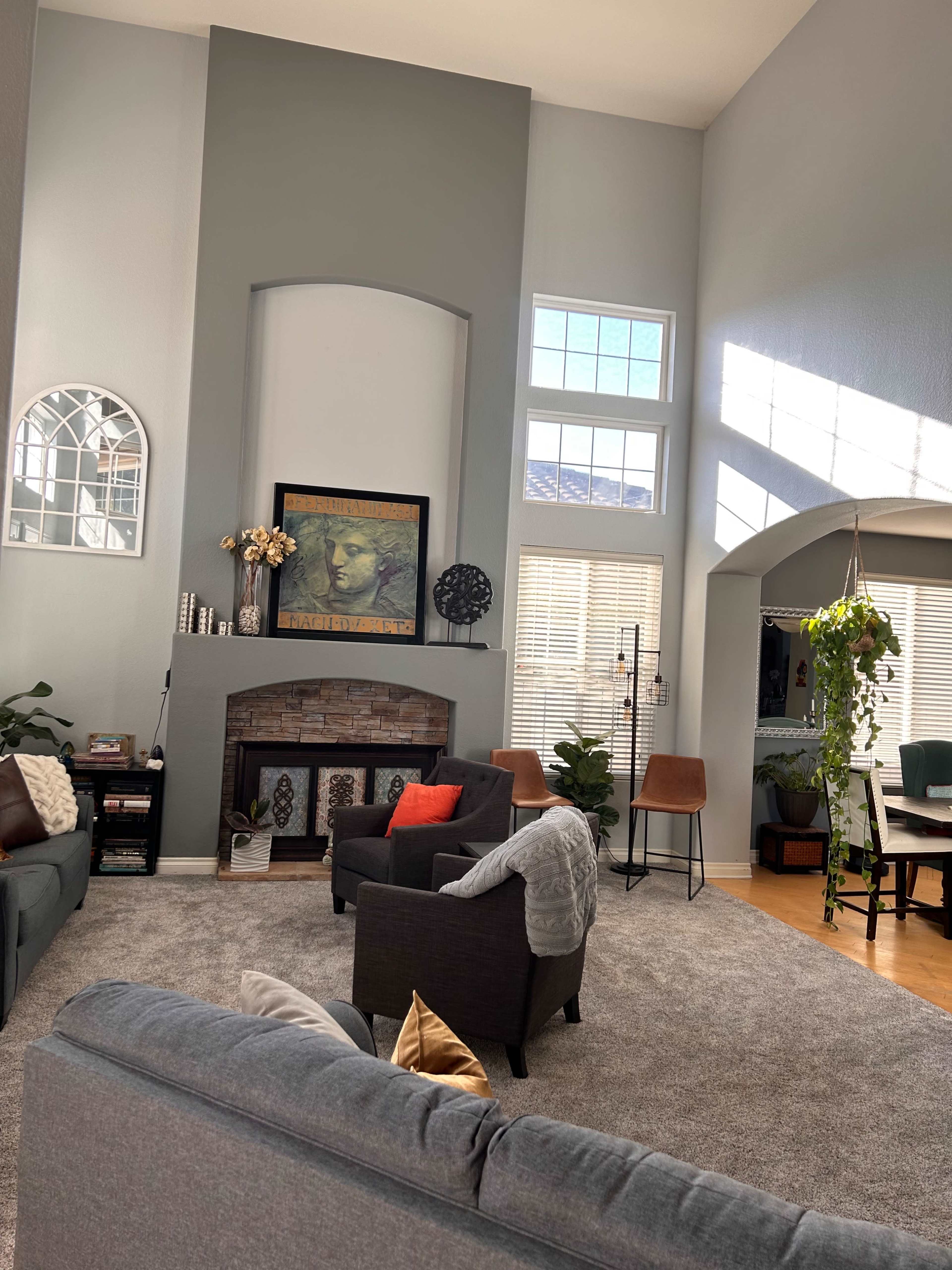 A spacious living room with high ceilings, featuring a large window that allows natural light in, and includes comfortable seating, a fireplace, and decorative plants.