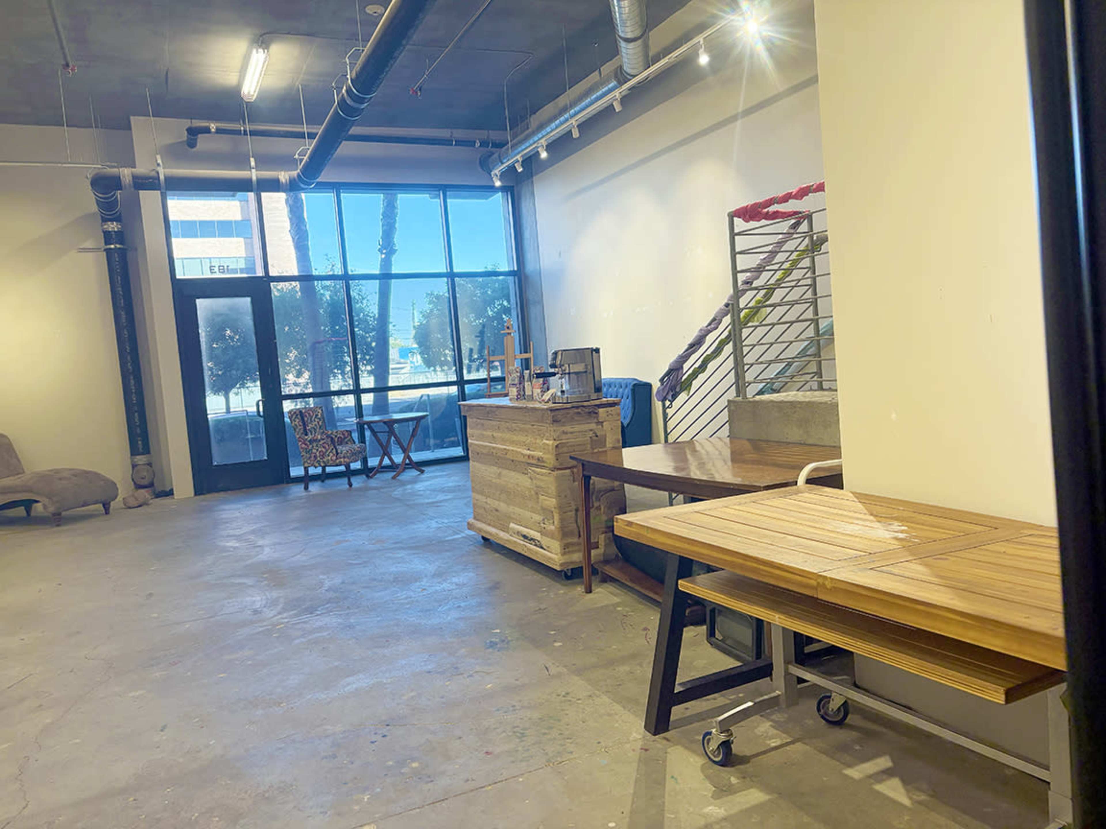 The image shows a spacious room with concrete flooring, featuring wooden furniture, large windows, and a staircase in the background.