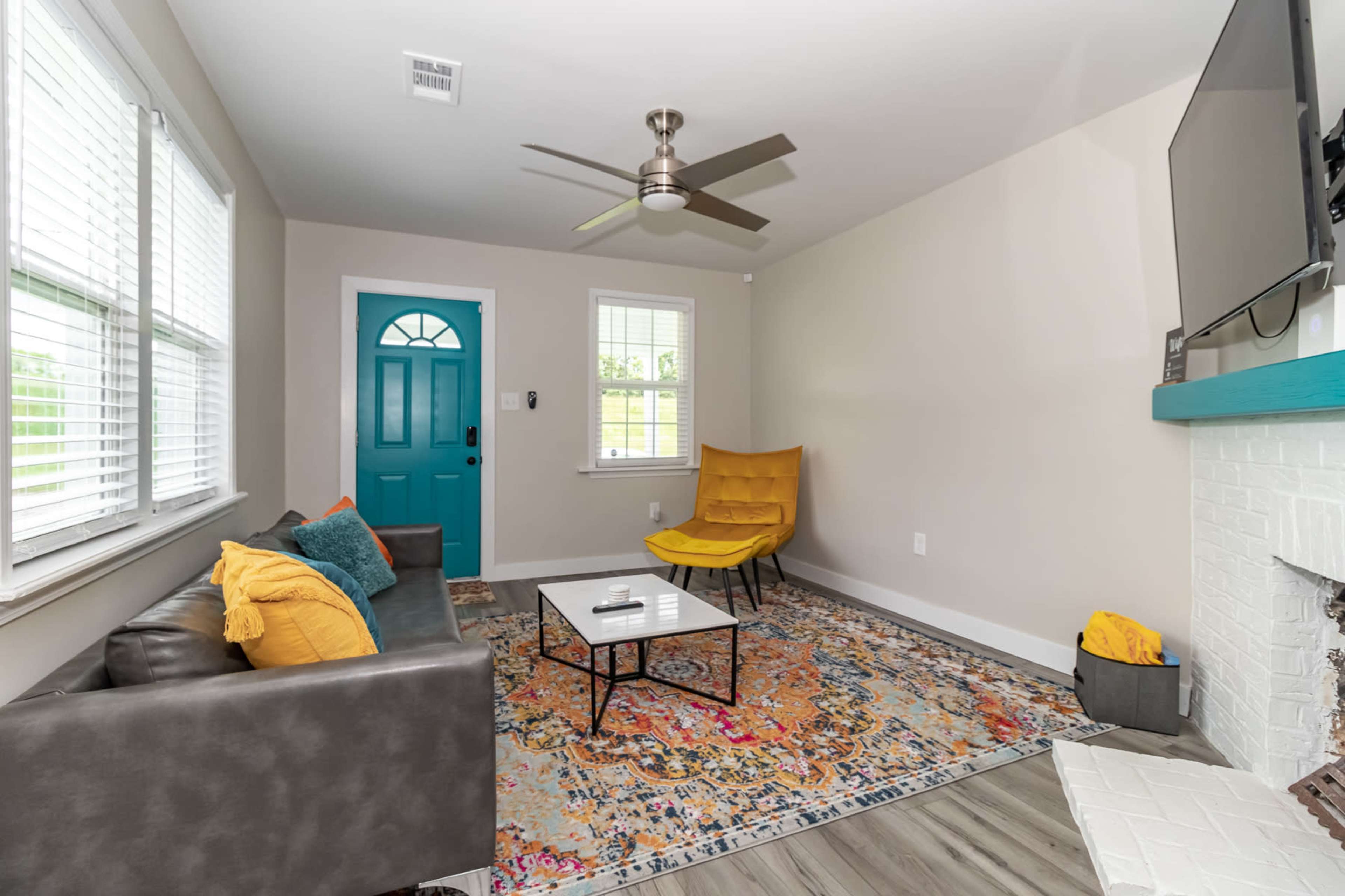 A living room with a teal front door, a gray sofa, a colorful rug, a coffee table, and two yellow accent chairs.