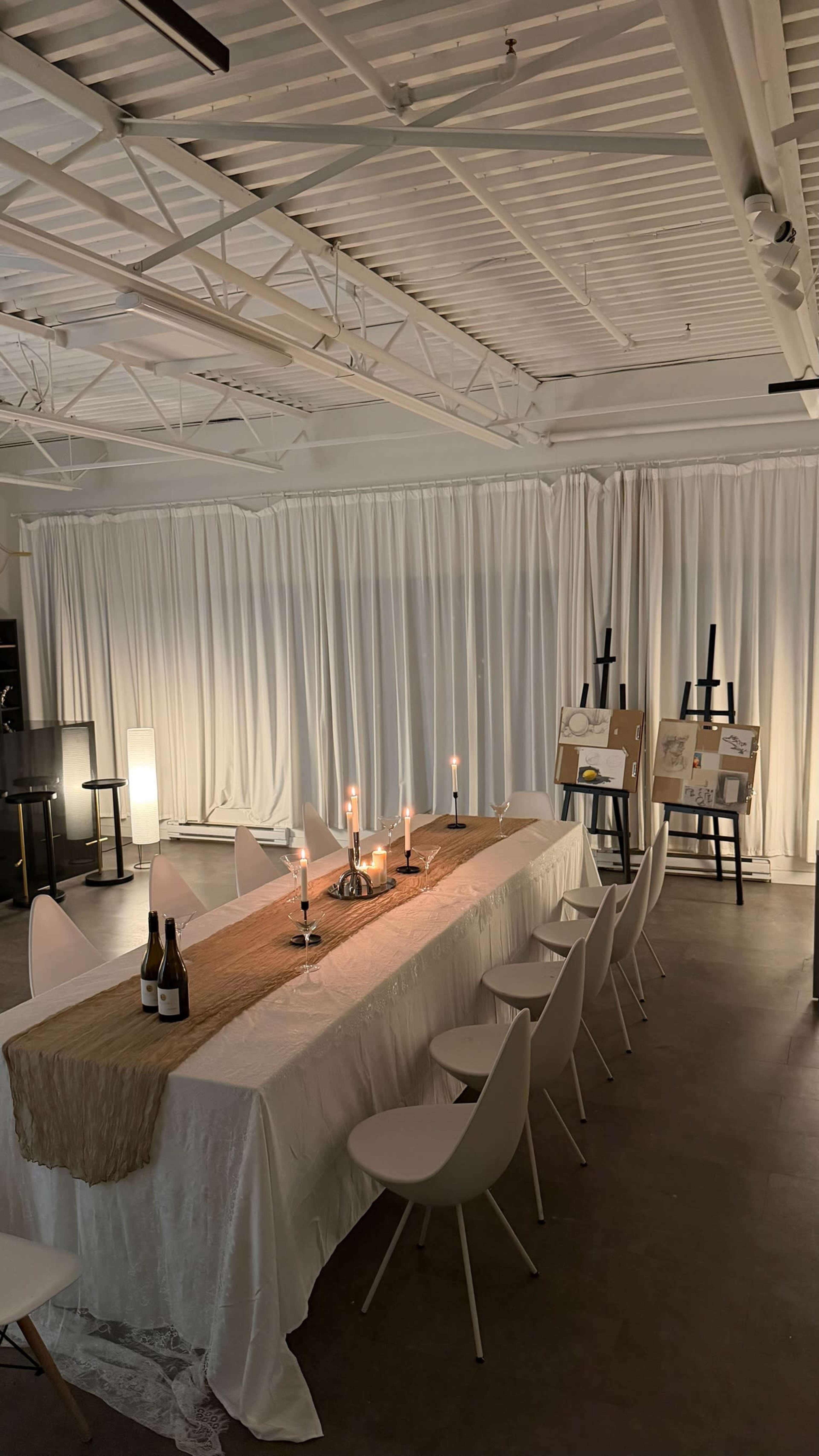 The image shows a long dining table set with candles and a tablecloth, surrounded by white chairs in a room with soft lighting and draped white curtains.