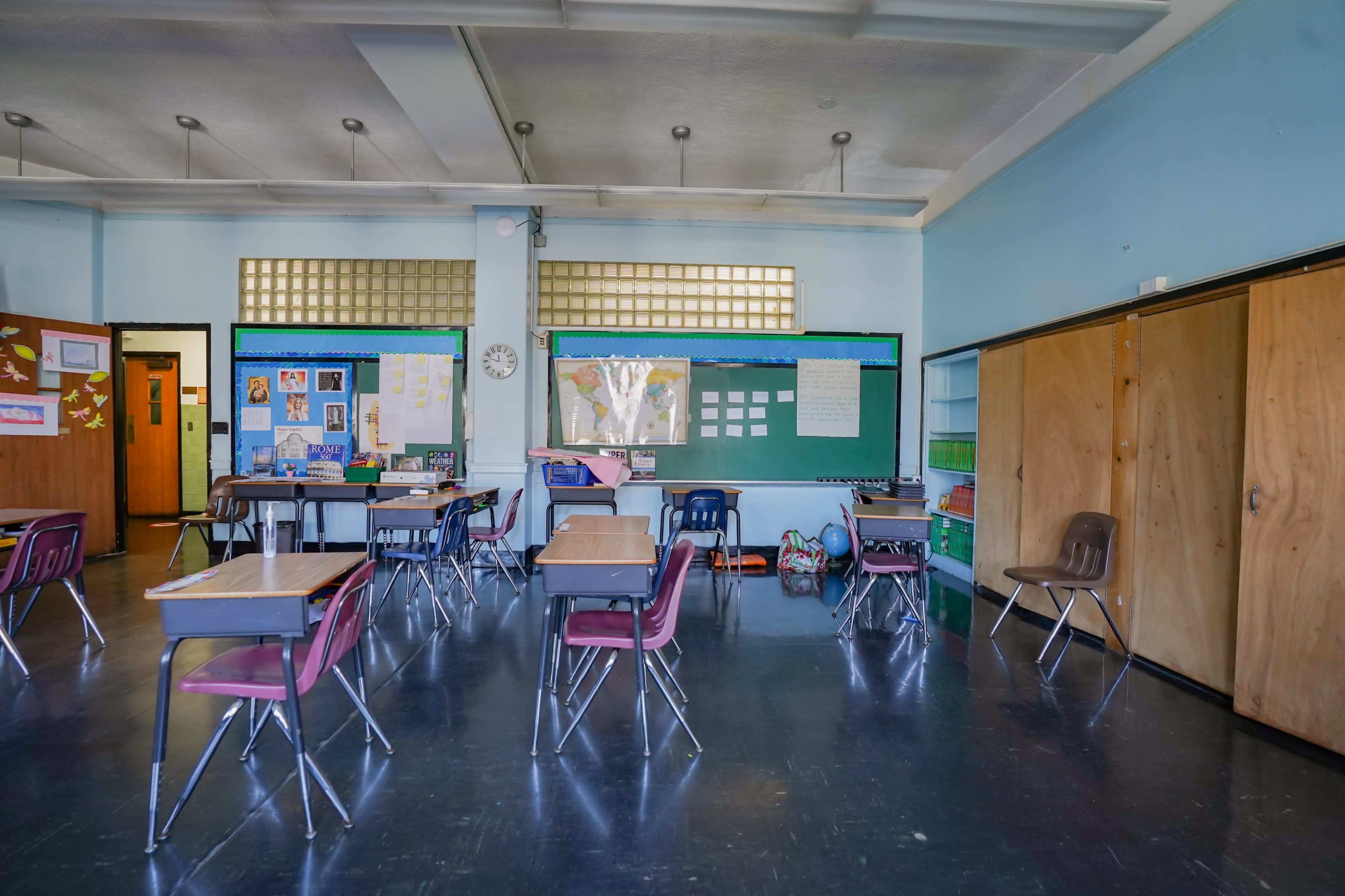 A classroom is arranged with several desks and chairs, featuring a chalkboard and bulletin boards on the walls.