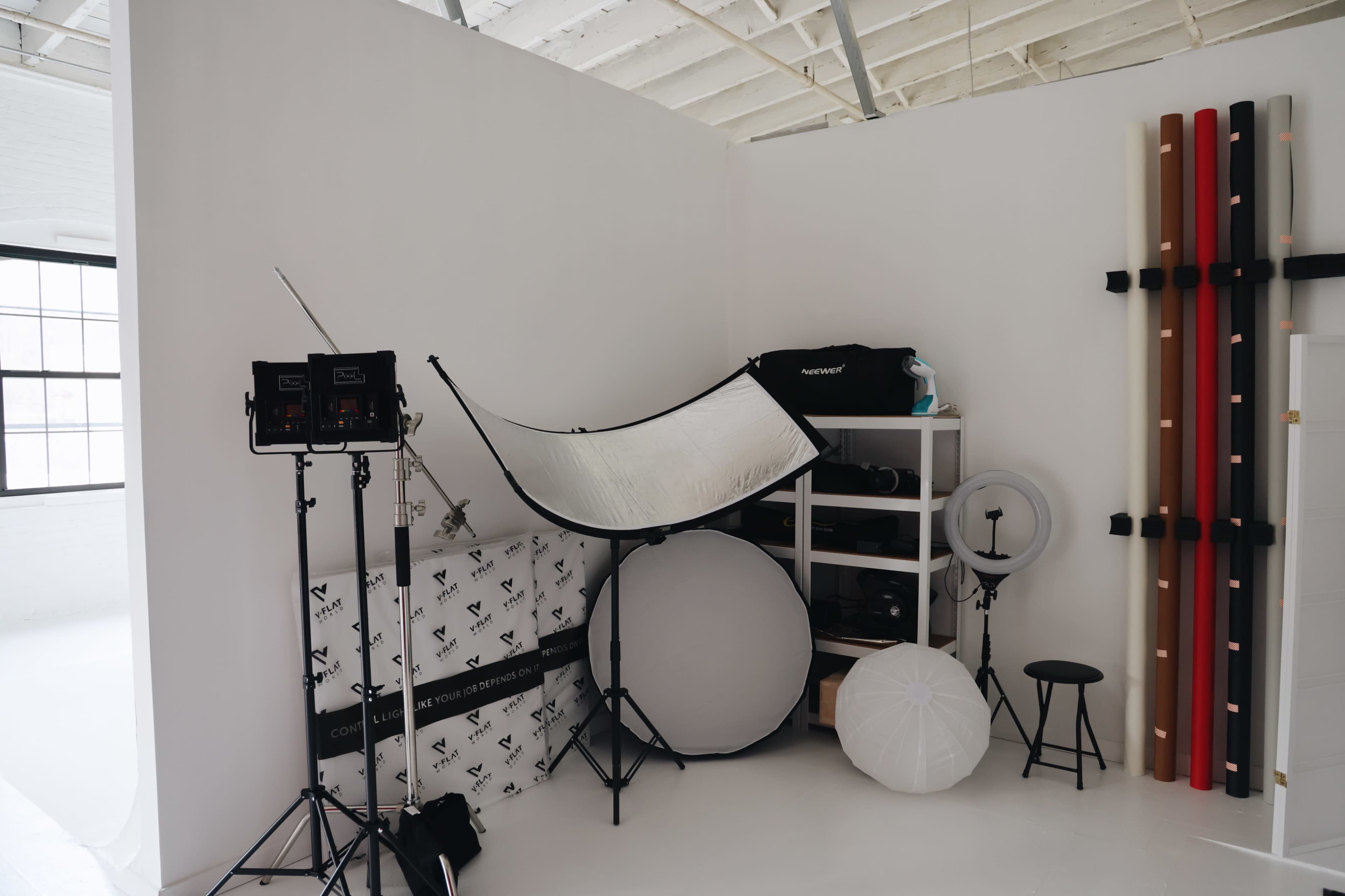 A photography studio corner with lighting equipment, reflectors, and a shelving unit filled with camera gear.
