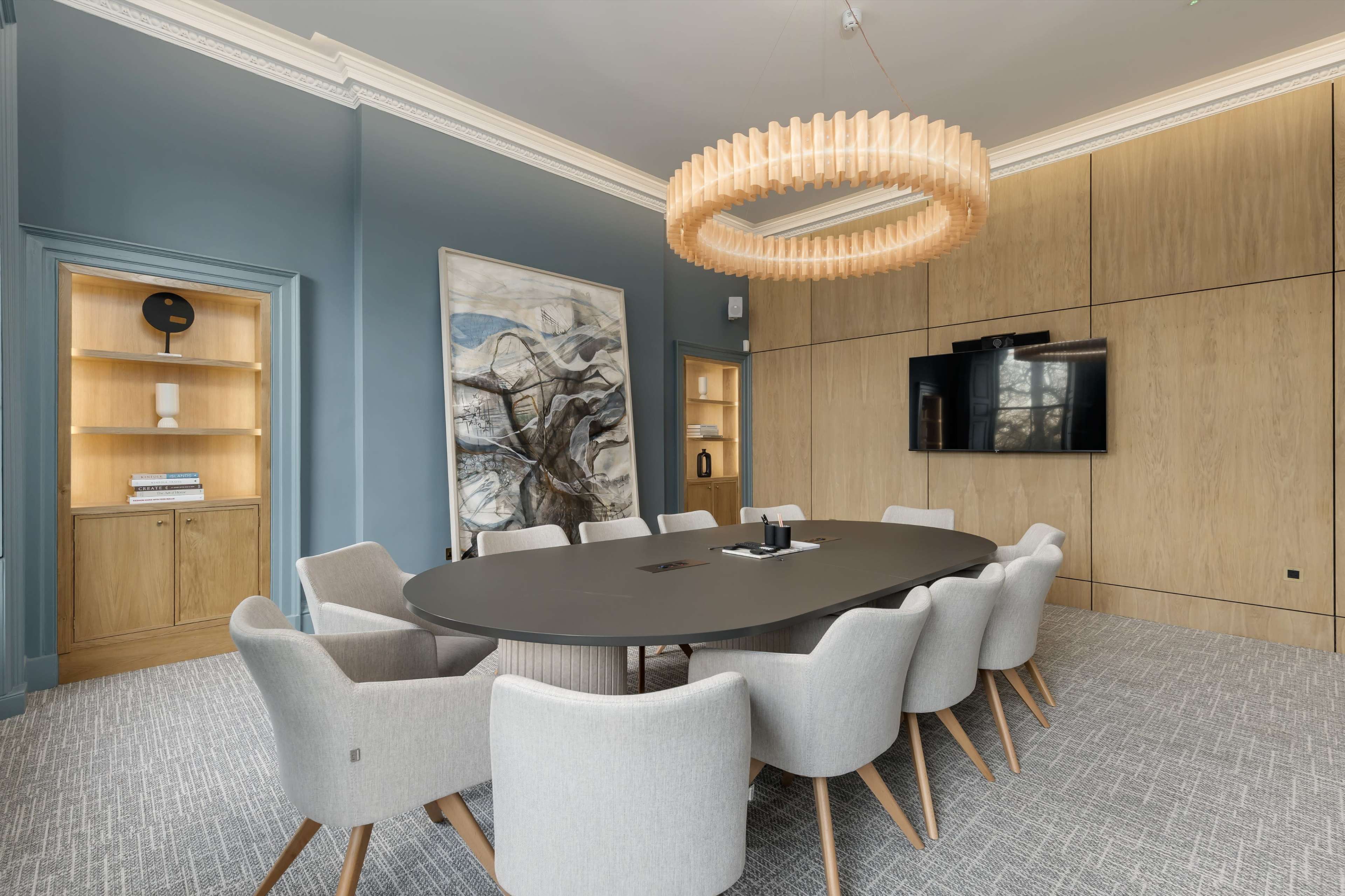 The image shows a modern conference room with a large oval table surrounded by ten gray upholstered chairs, featuring a wooden wall and a large light fixture above.
