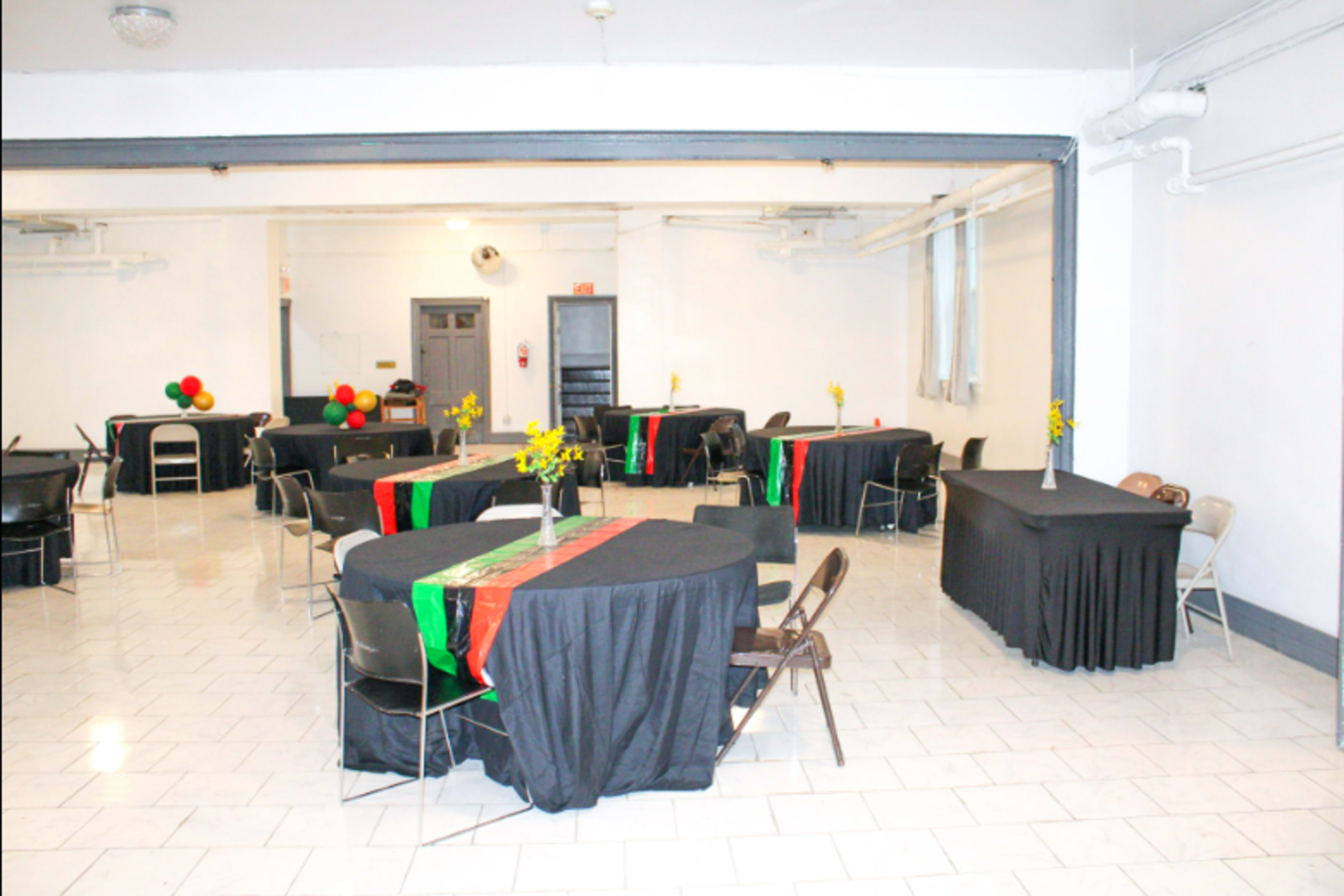 The image shows a spacious event room with several round tables covered in black tablecloths, each adorned with colorful centerpieces and decorations.