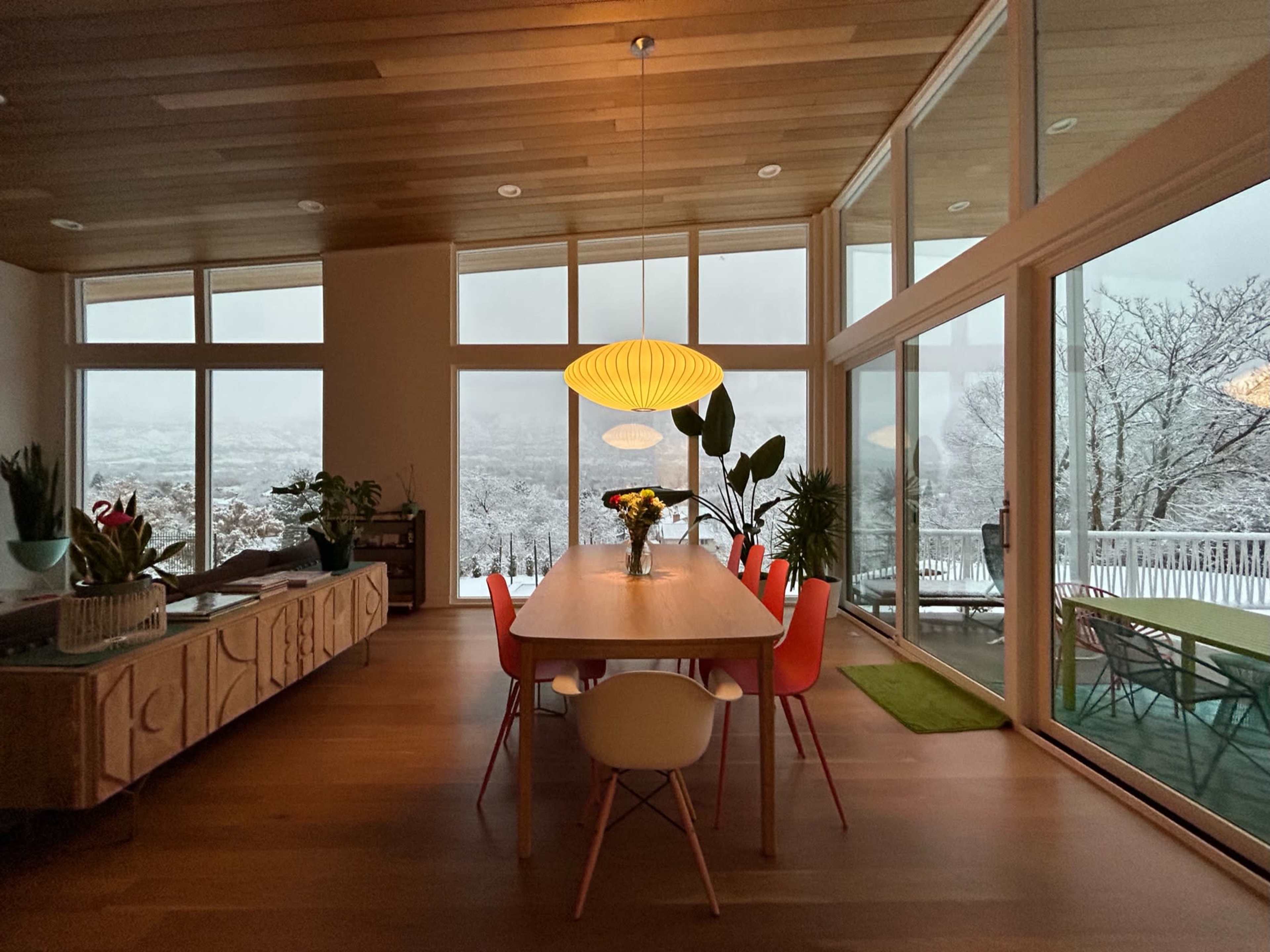 A modern dining area features a long table surrounded by colorful chairs, with large windows showcasing a snowy landscape outside.