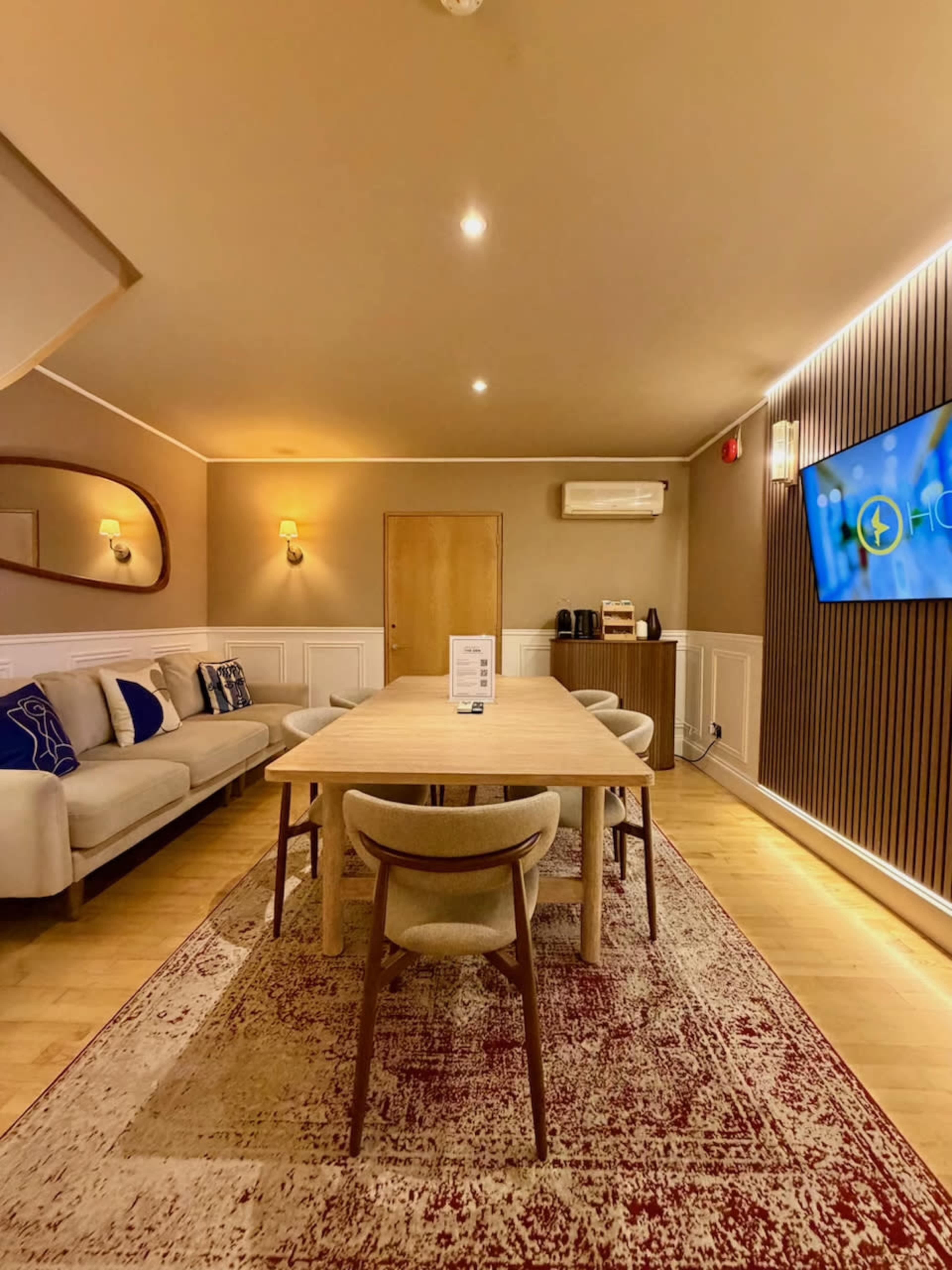 The image shows a modern meeting room featuring a long wooden table, beige chairs, a sofa, and a wall-mounted television.