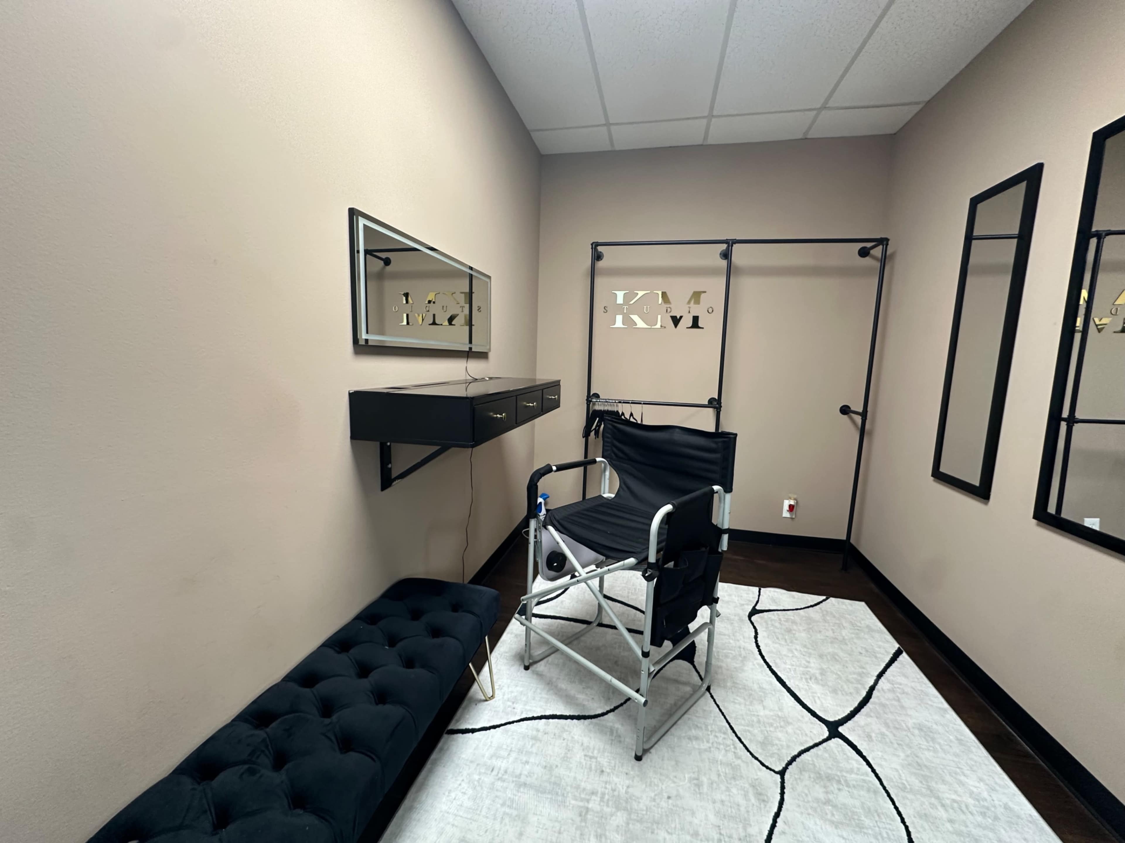 The image shows a small room with a makeup or styling chair, a wall-mounted mirror, a black storage unit, and a decorative area rug.