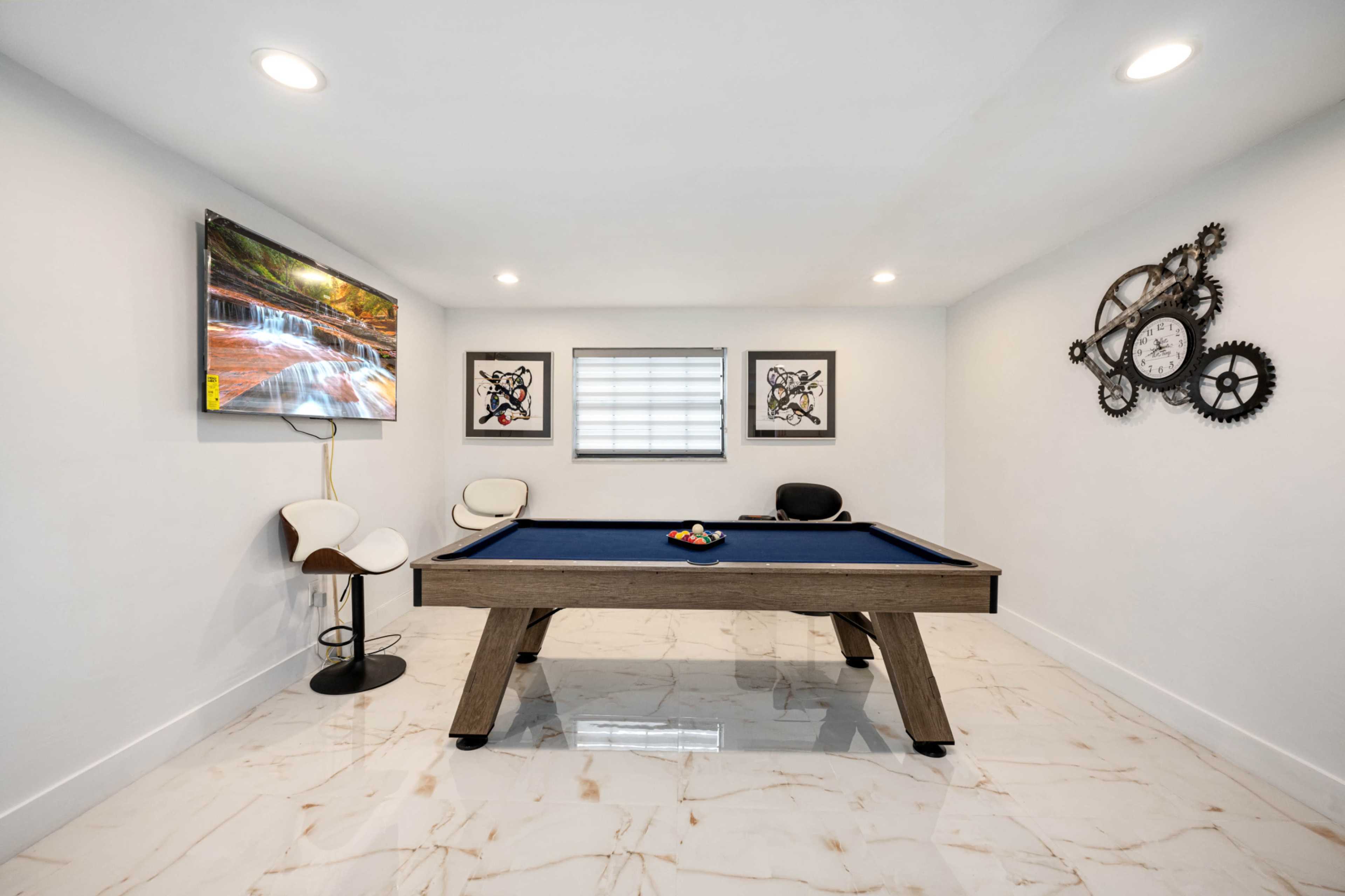 The image shows a modern game room featuring a pool table, a television mounted on the wall, and decorative artwork.