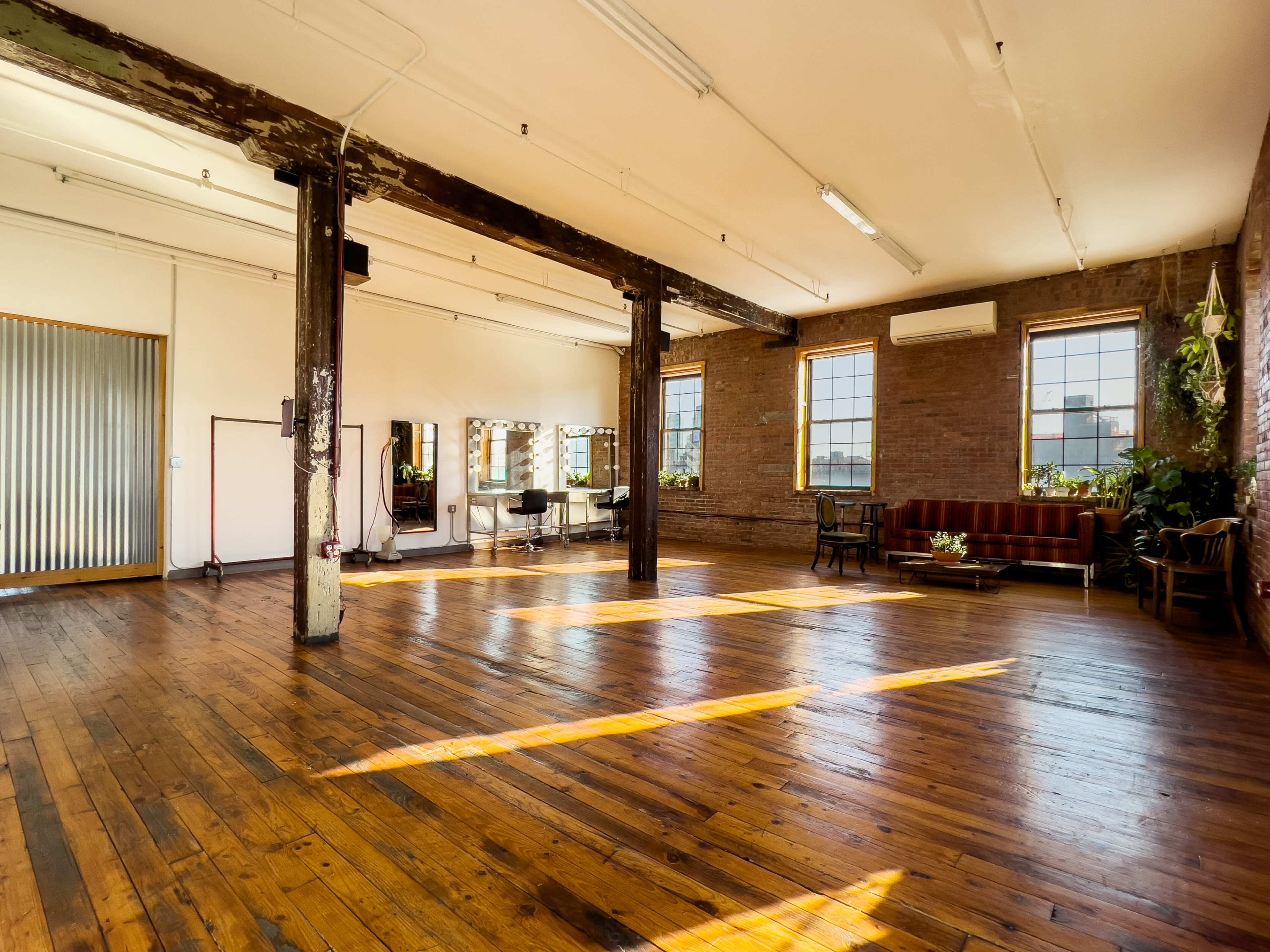 A spacious, well-lit room with wooden floors, brick walls, and large windows, featuring a few furniture pieces and mirrors.