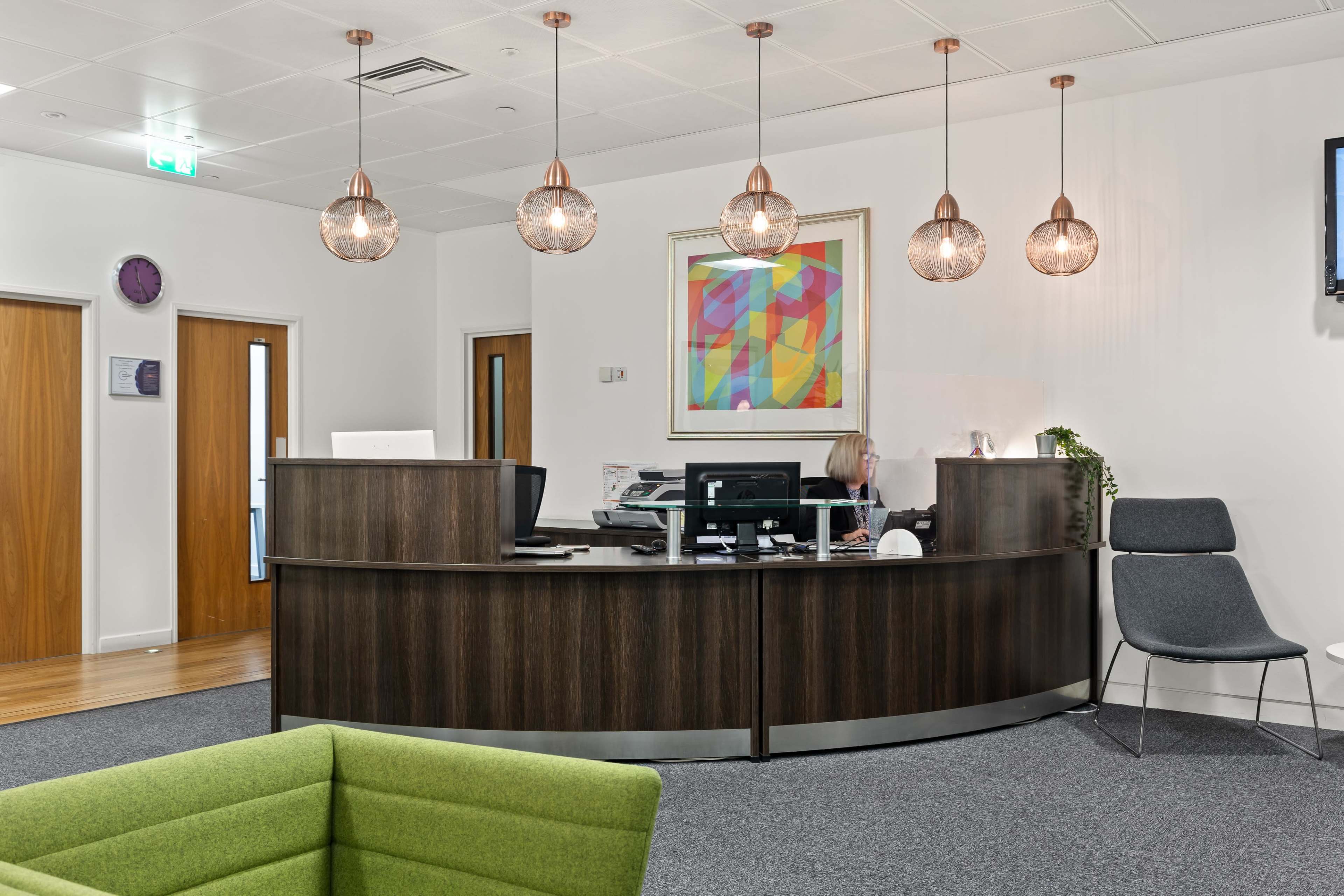 A modern reception area with a wooden curved desk, a computer, and a seated receptionist, surrounded by decorative lighting and a colorful artwork.