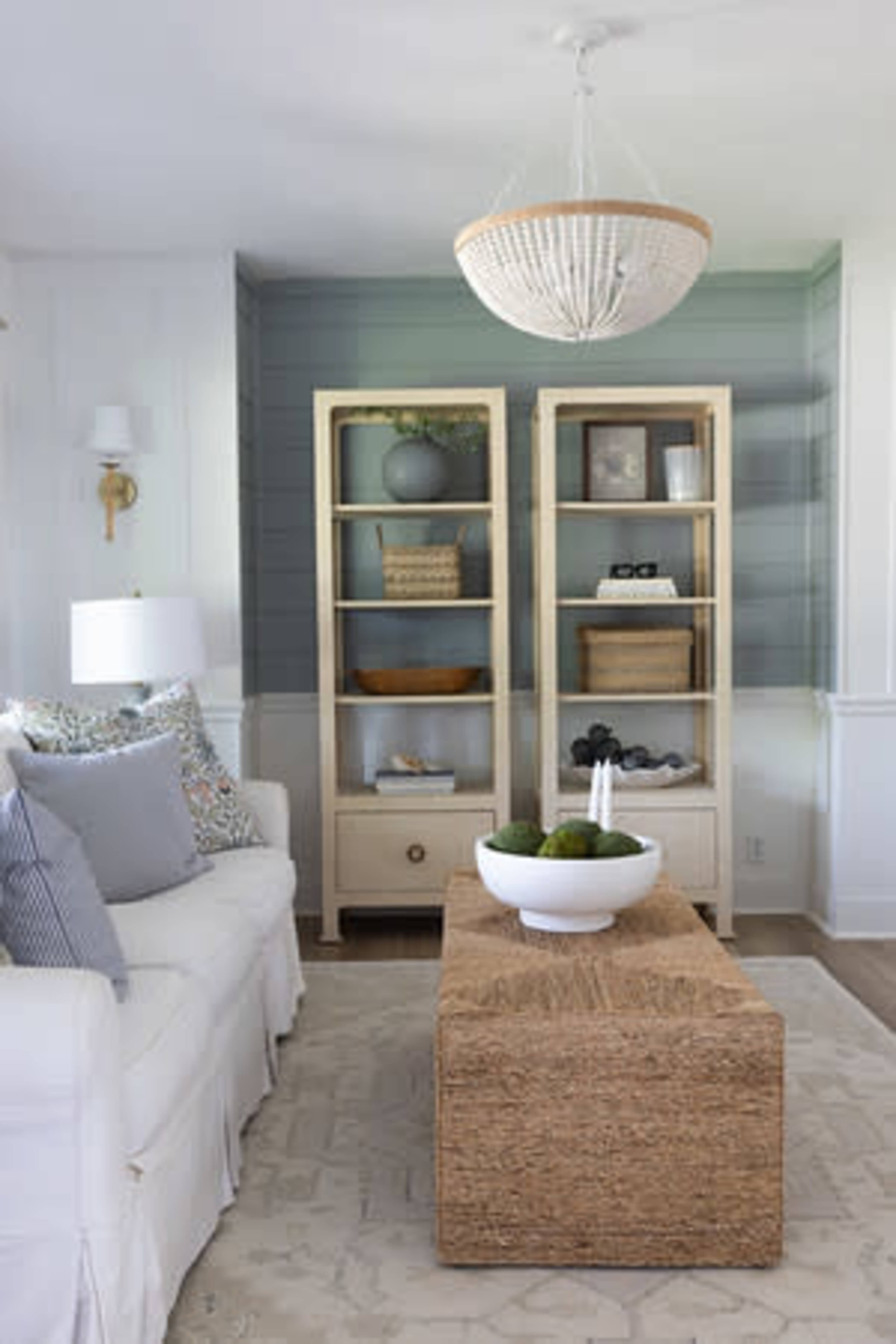 The image depicts a bright living room featuring a white sofa with decorative pillows, a natural woven coffee table, and two open bookshelves flanking a light fixture overhead.