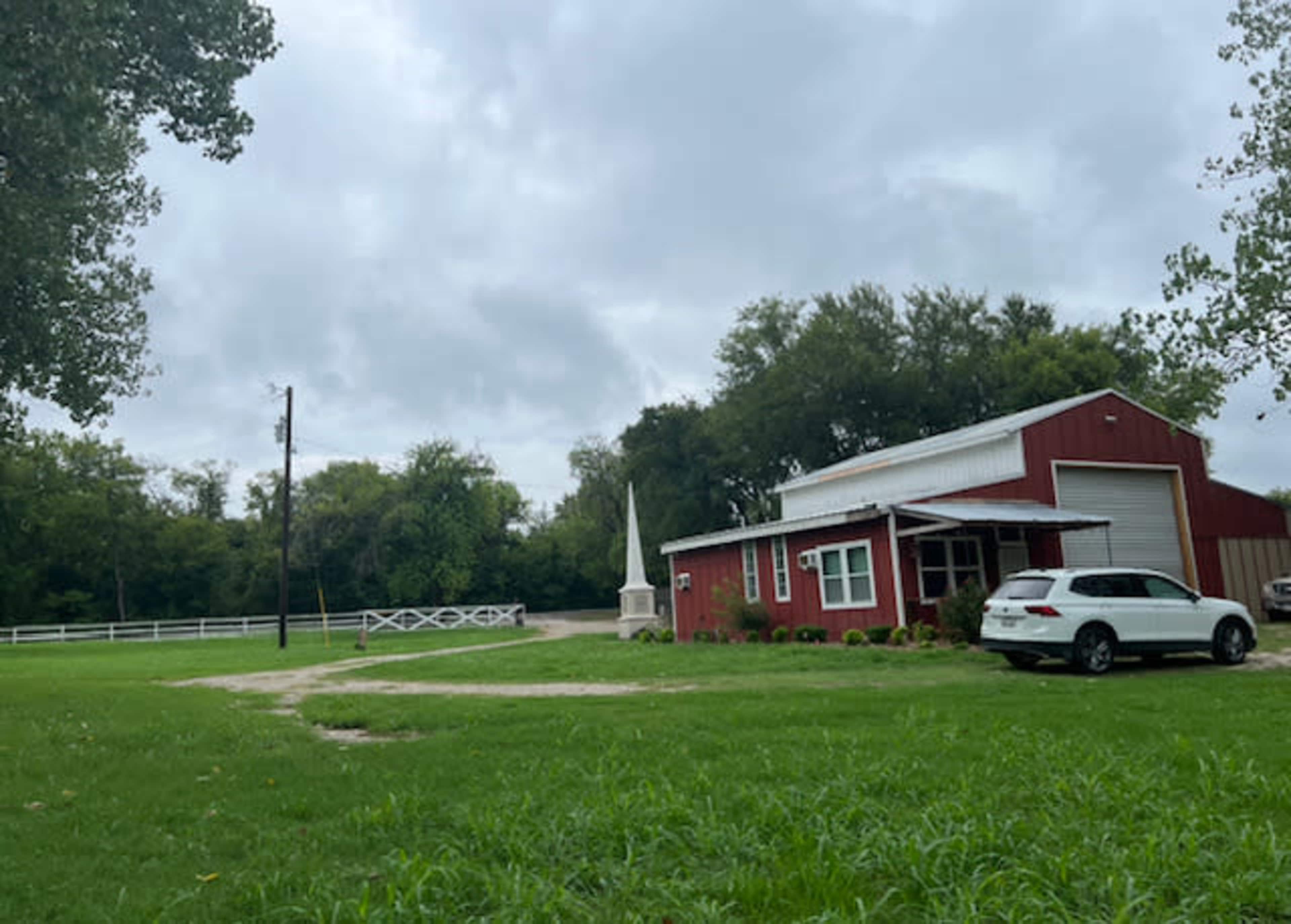 A red barn-like building with a white vehicle parked nearby is situated on a grassy area beside a dirt path, with trees and a white fence in the background.