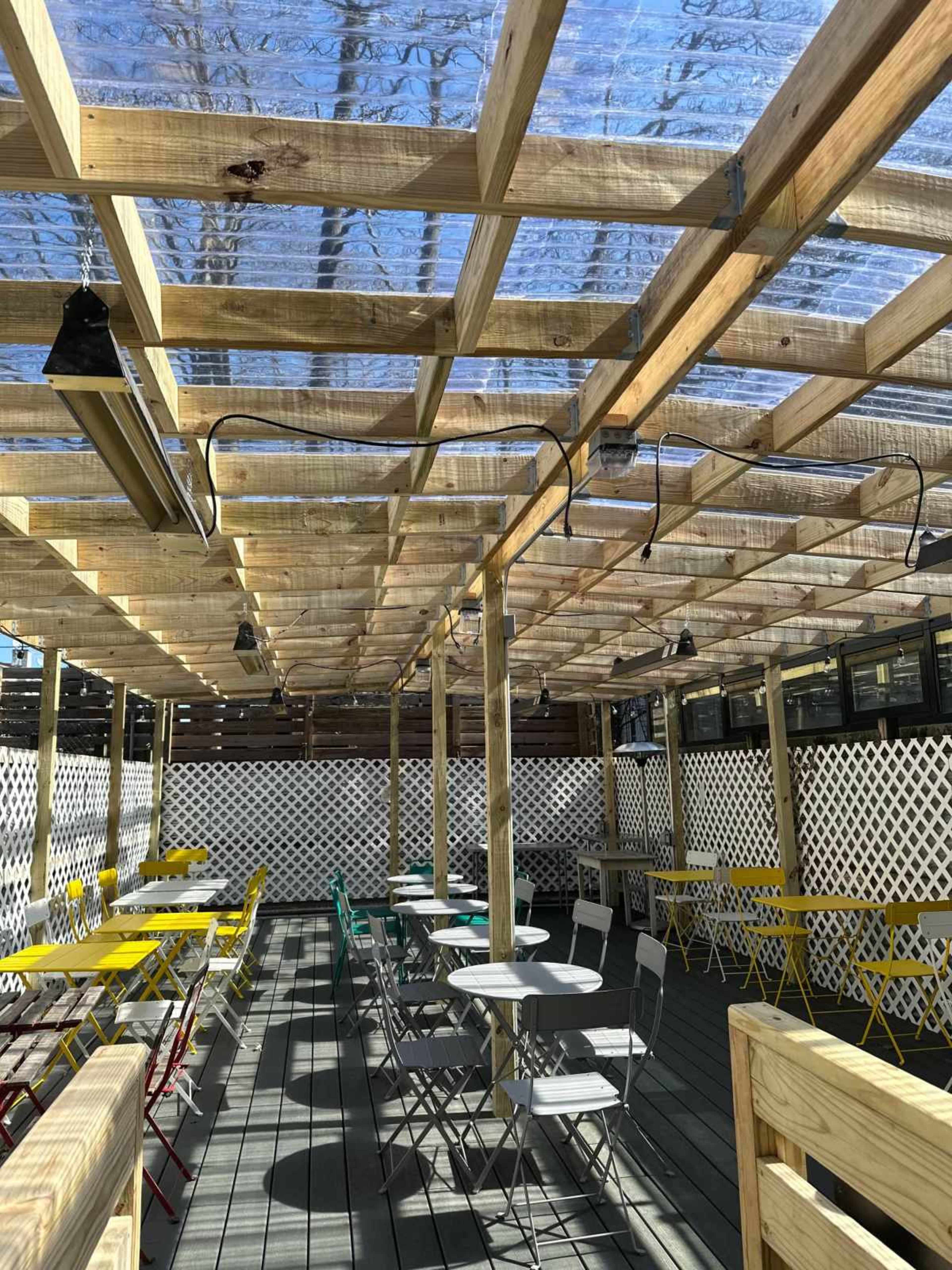 The image shows a shaded outdoor dining area with wooden beams and various colorful chairs and tables arranged on a wooden deck.