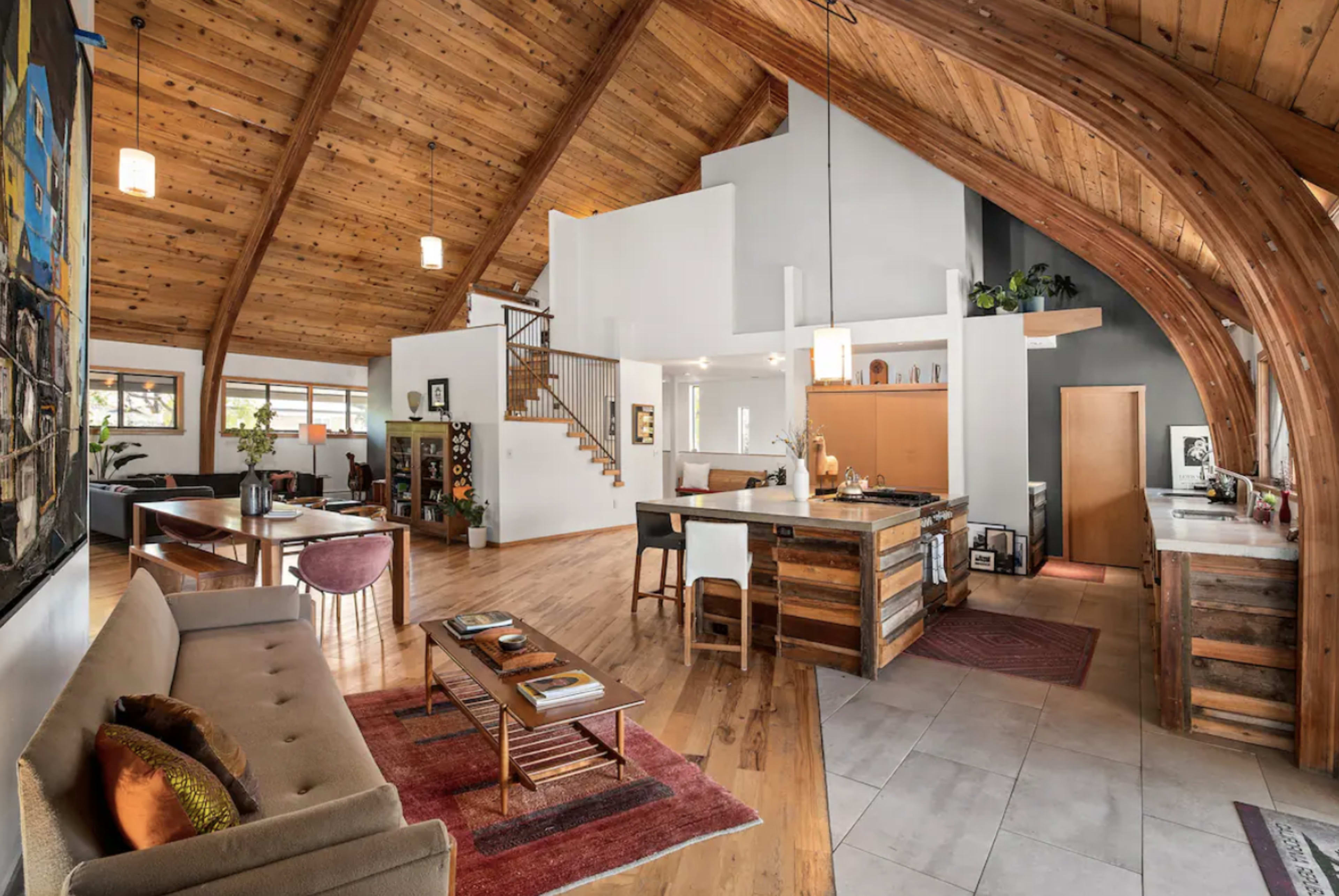 A spacious, modern living area with wooden beams, a staircase, and an open kitchen featuring a large island and dining area.