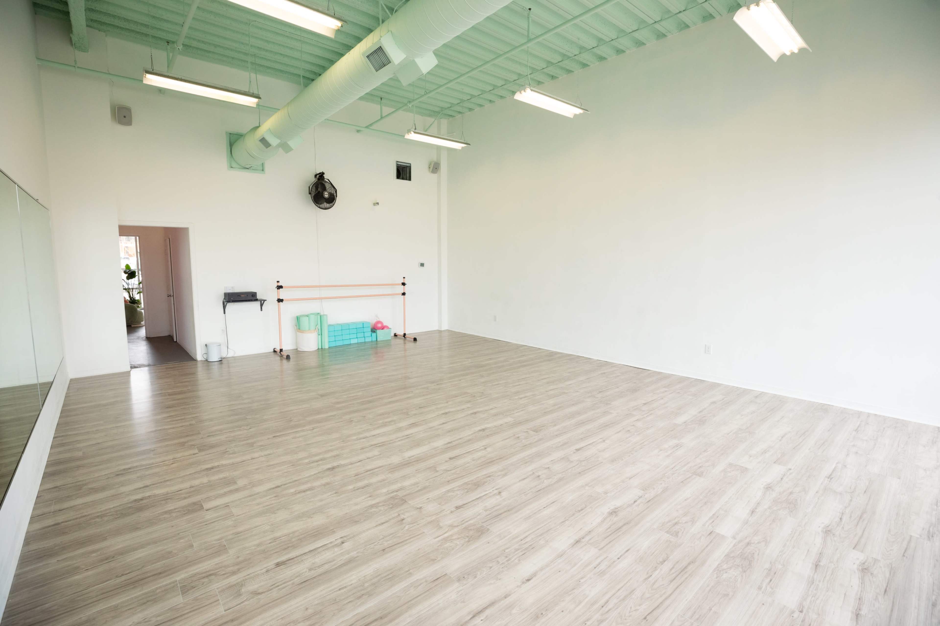 A spacious, empty room with wooden flooring, large mirrors on one wall, and minimal furniture, including a barre and some exercise equipment.