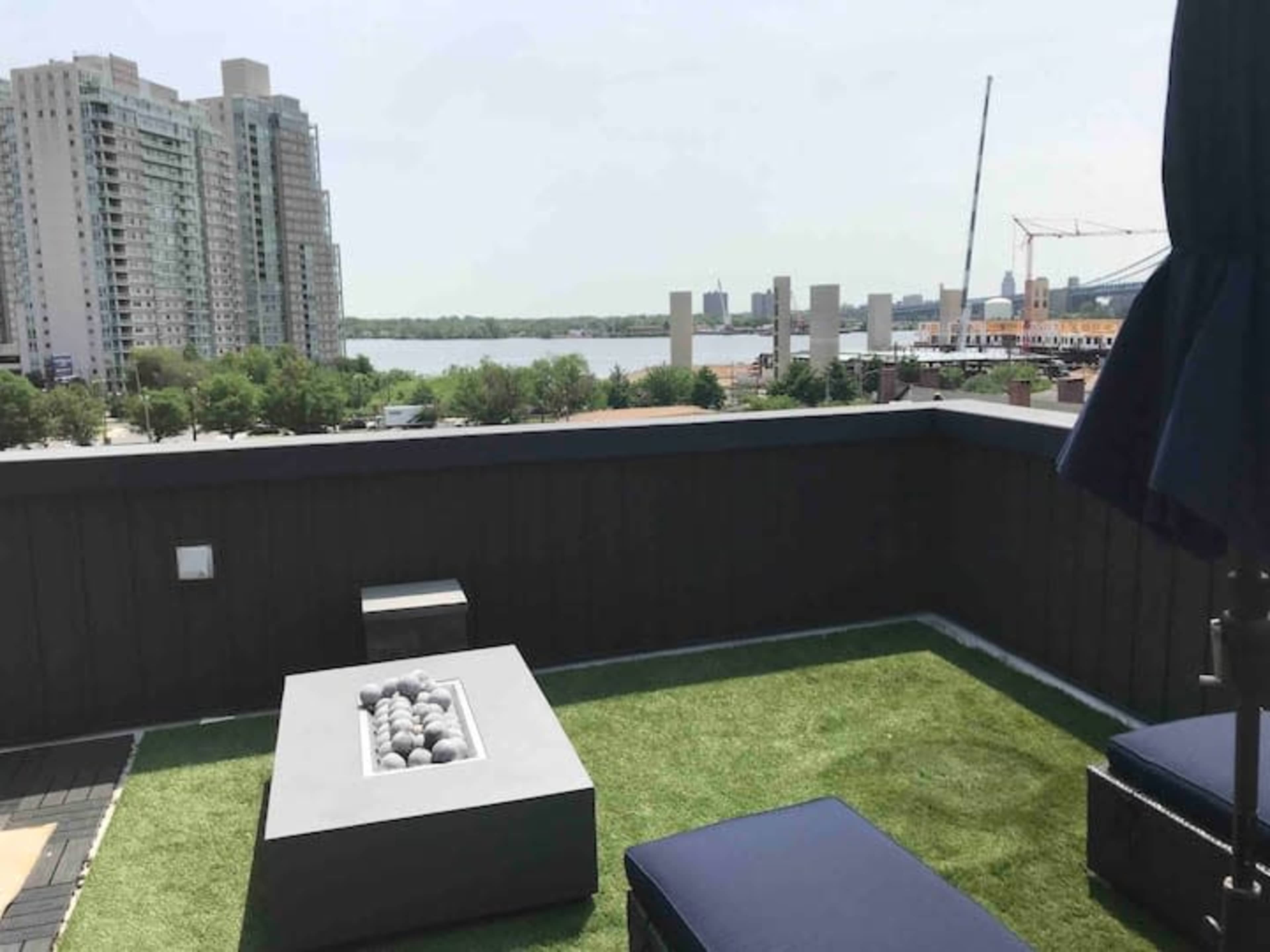 A rooftop patio with a fire pit surrounded by green grass and modern furniture, overlooking a body of water and buildings in the distance.