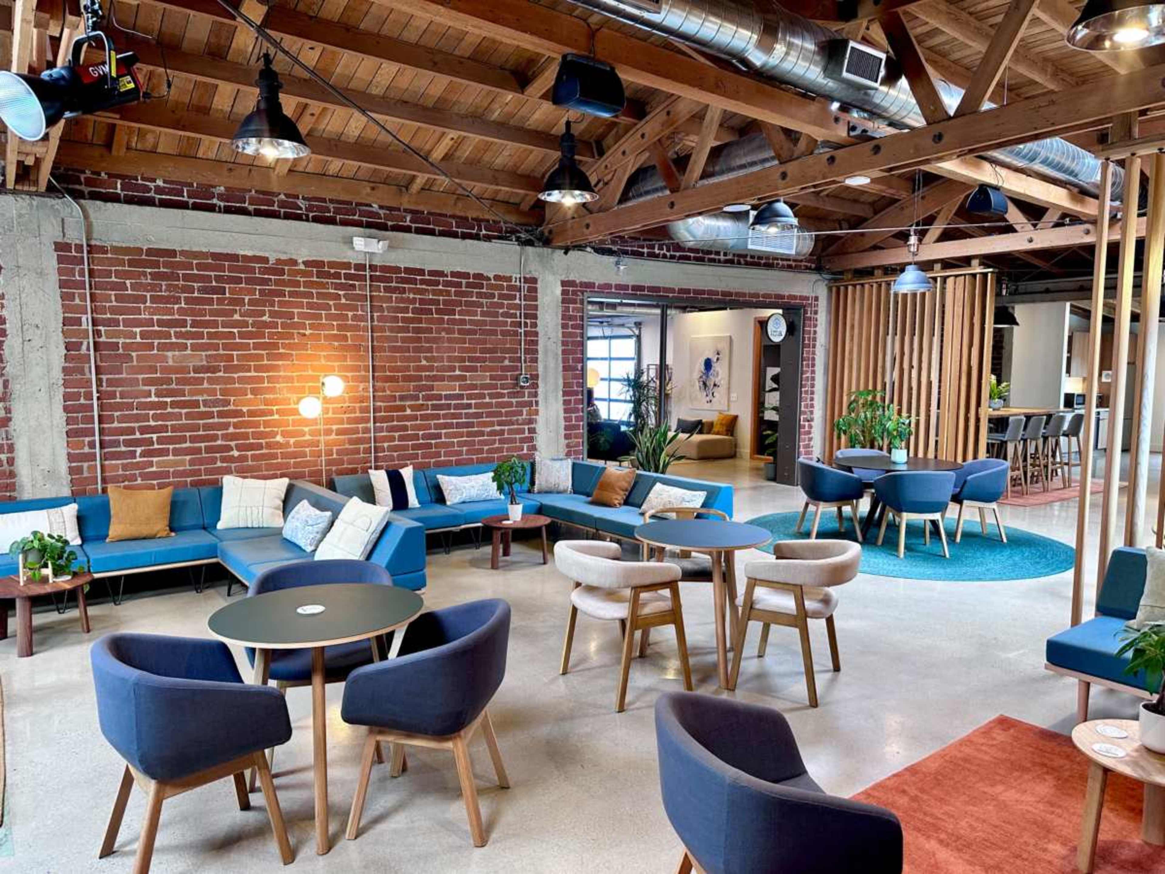 The image shows a modern lounge area with sectional sofas, circular tables, and wooden accents, featuring exposed brick walls and a high ceiling with industrial lighting.