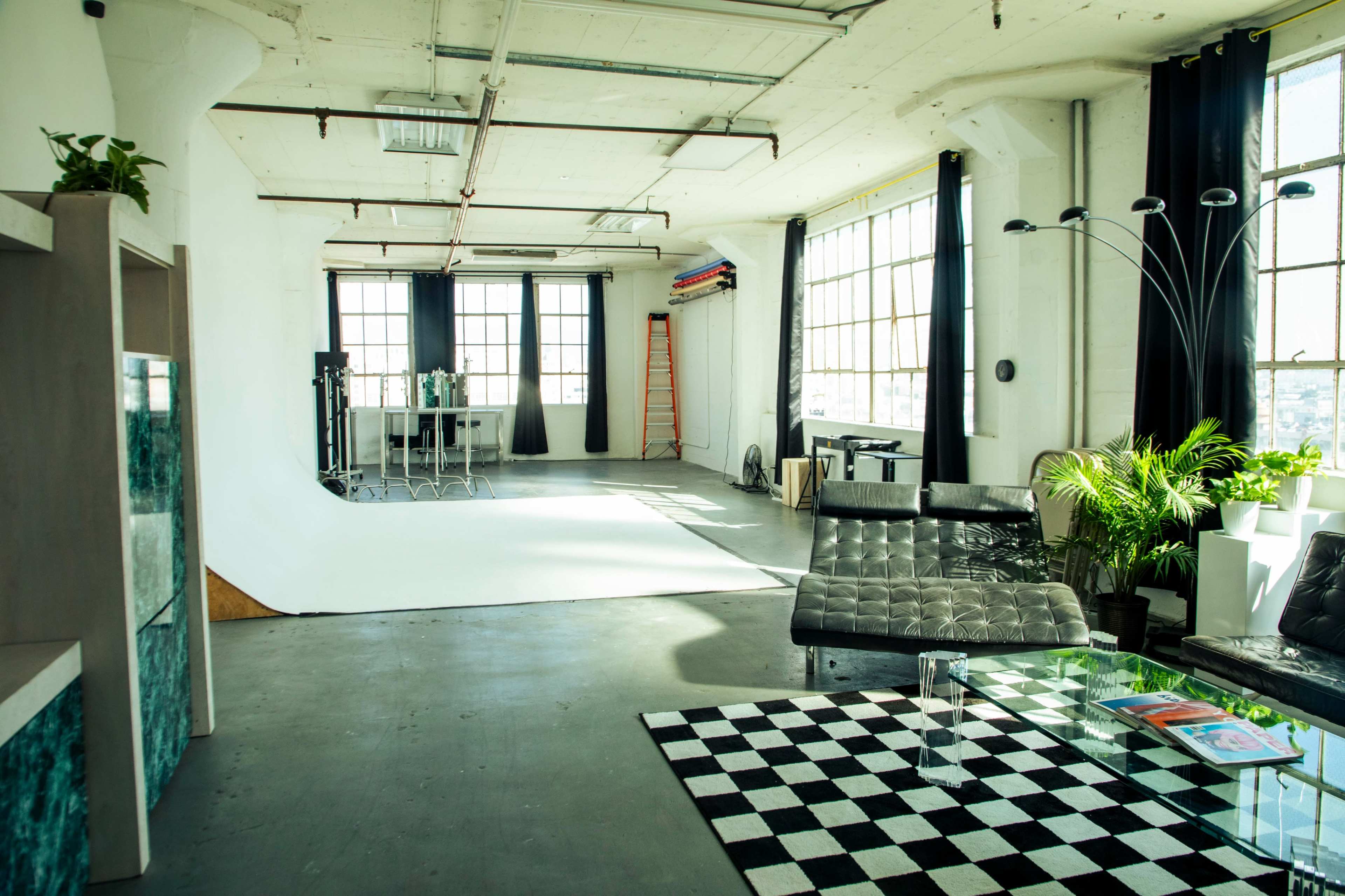 The image shows a spacious, well-lit studio with a large white backdrop, modern furniture, and a checkered rug.