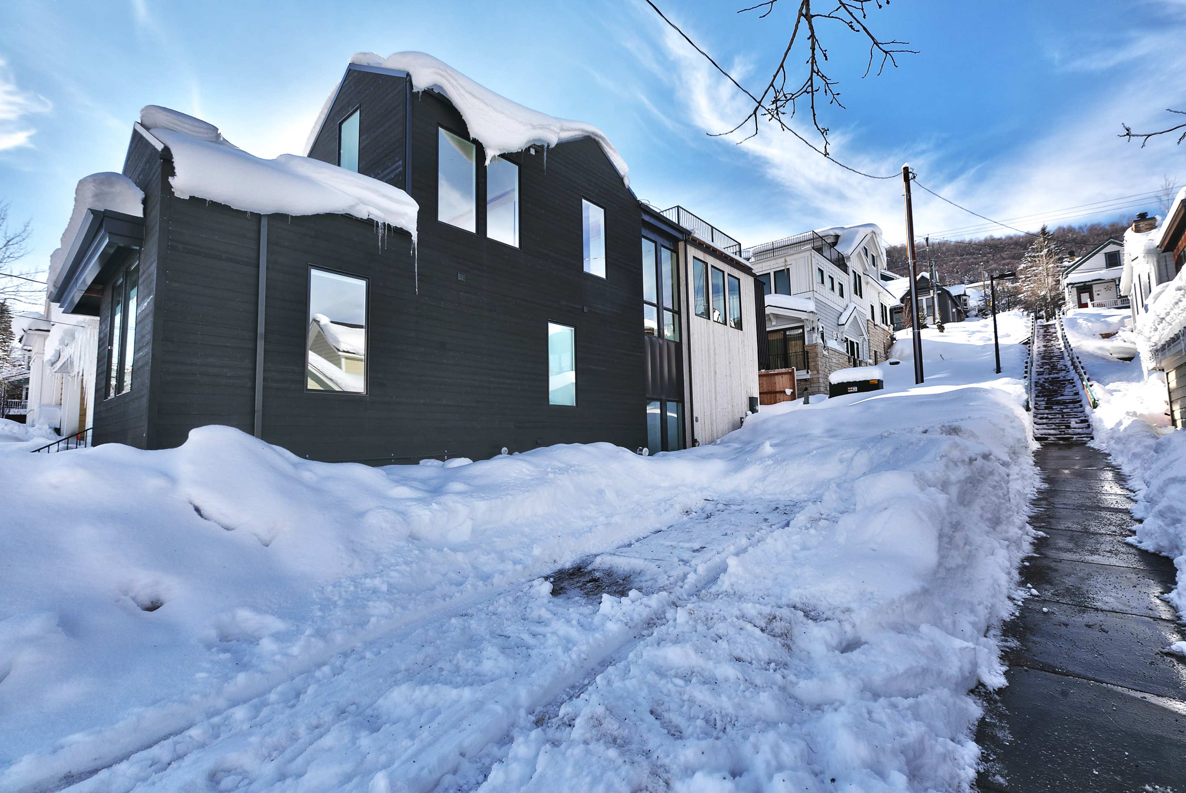 A snow-covered residential area features modern homes with large windows and a clear blue sky overhead.
