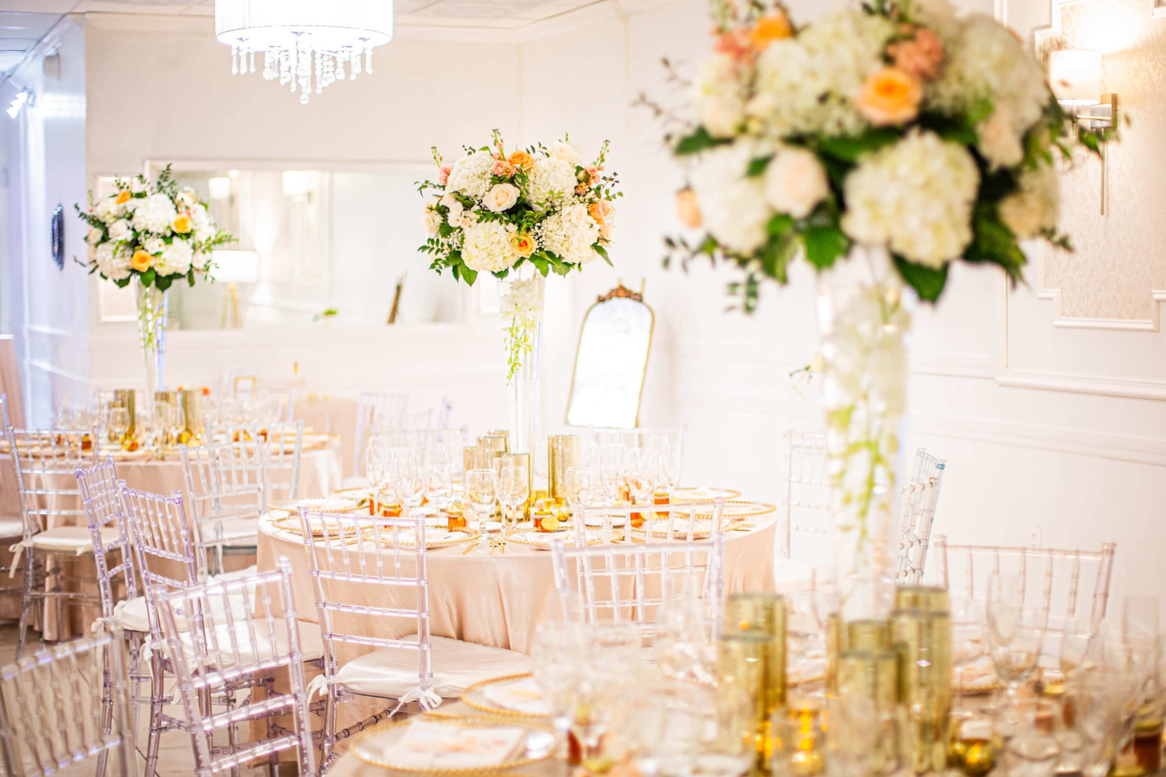 The image shows an elegantly decorated banquet hall with tall floral centerpieces on each table, featuring round tables set with glassware and gold accents.