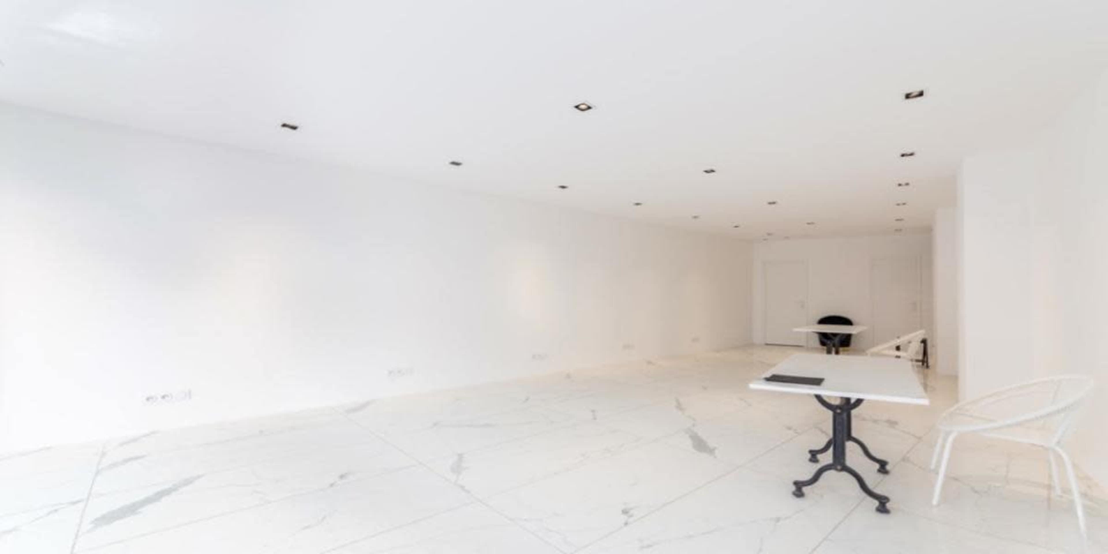 The image shows a spacious white room with marble flooring, featuring recessed lighting and a minimalistic table and chair setup.