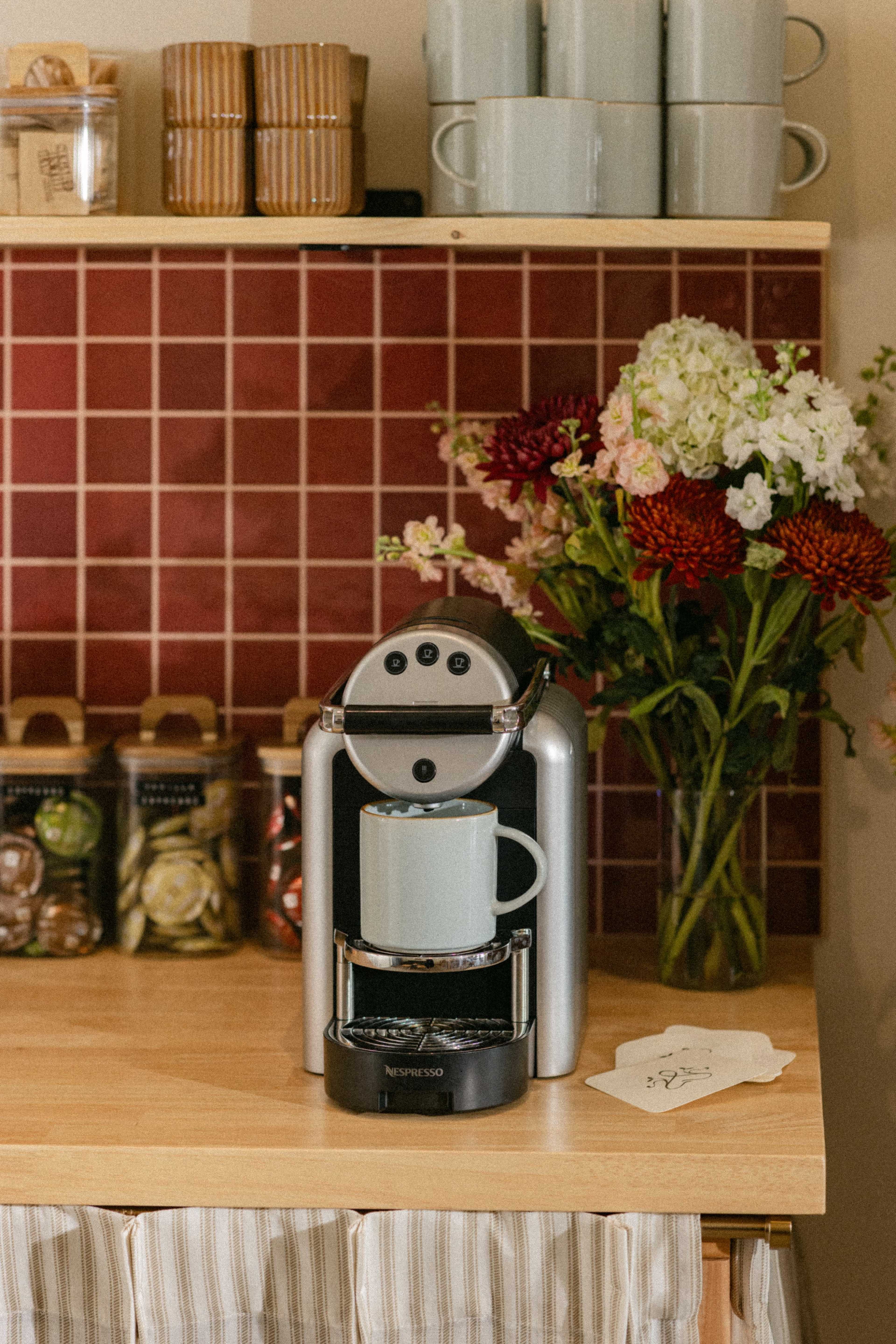 A Nespresso coffee maker sits on a wooden countertop with a light blue mug, surrounded by jars of food items and a vase of colorful flowers.