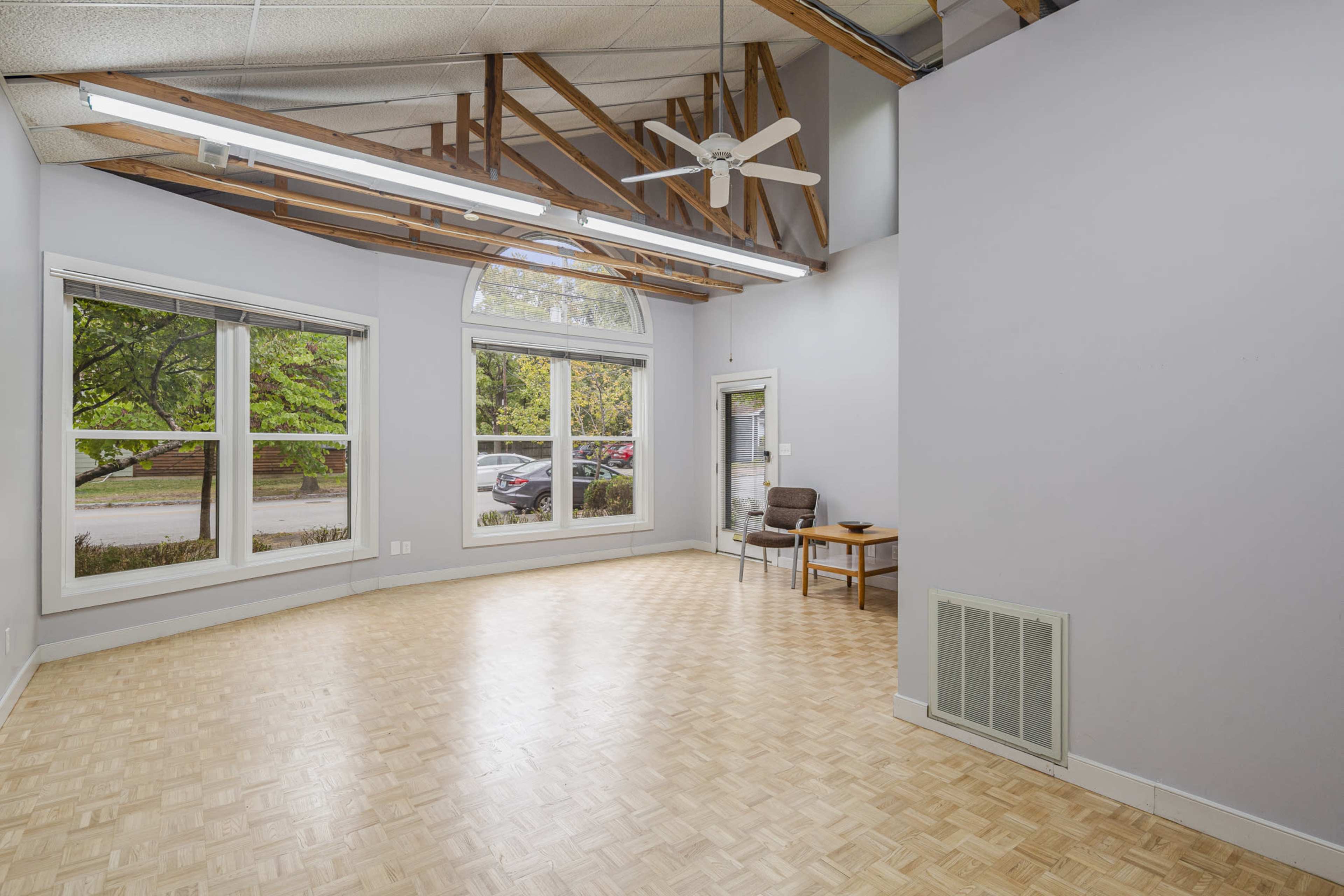 A spacious room with wooden flooring, large windows, and a ceiling fan, featuring minimal furniture.