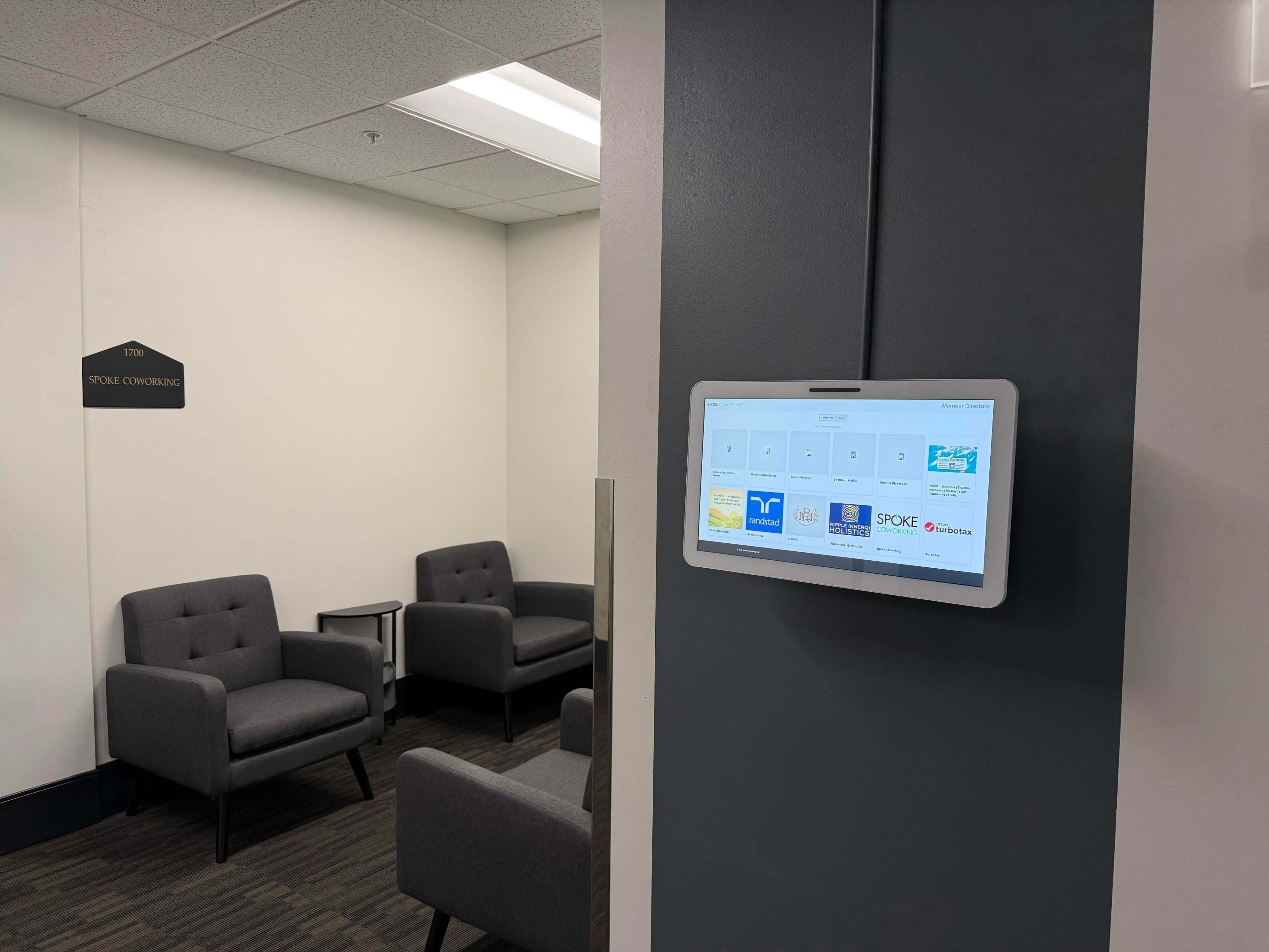 A waiting area features two gray armchairs and a wall-mounted digital display showing meeting room options.