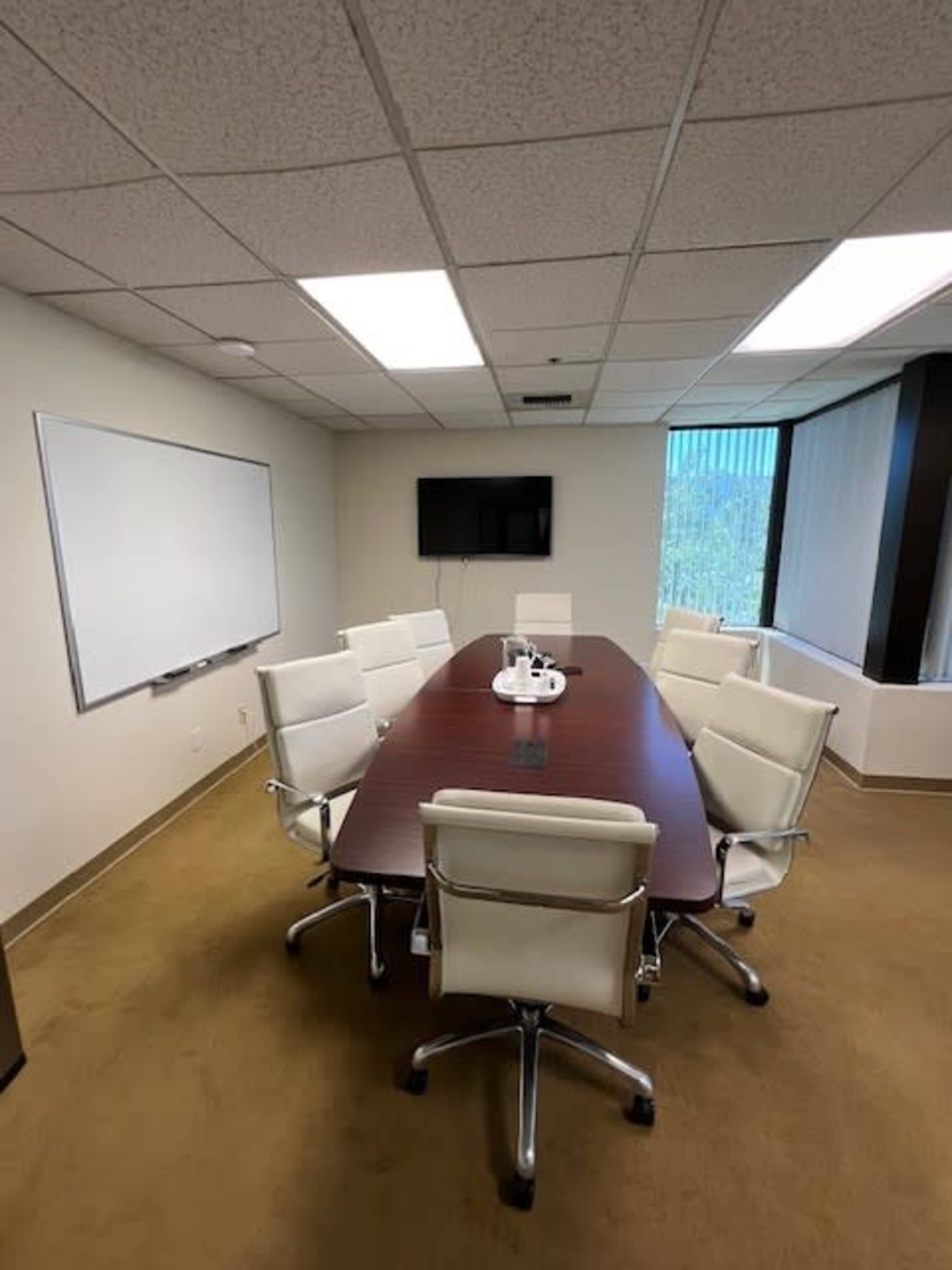A conference room with a large wooden table surrounded by eight white leather chairs, a whiteboard, and a wall-mounted TV.