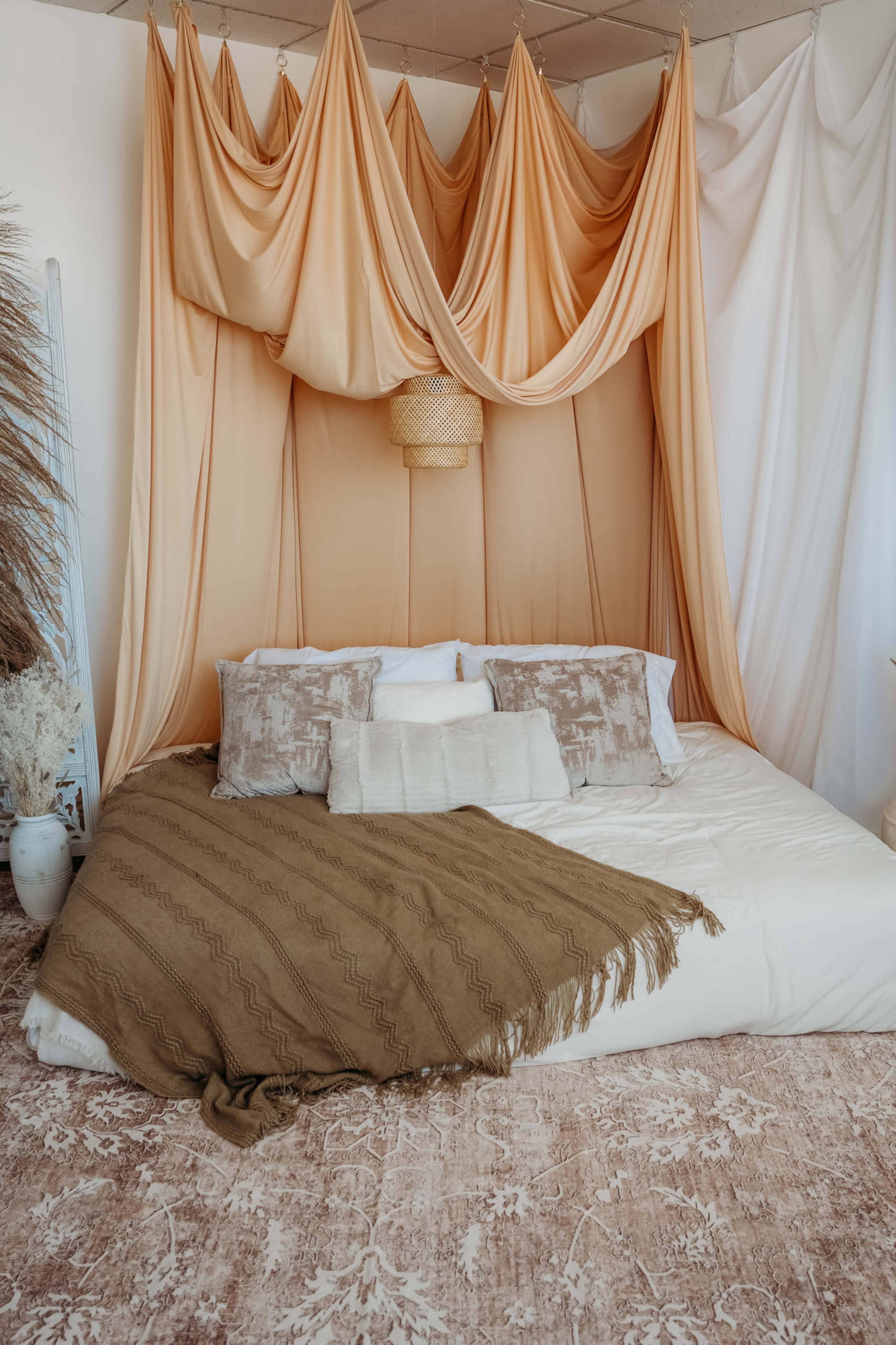 The image shows a cozy bedroom with a large bed adorned with decorative pillows and a soft brown blanket, set against a backdrop of flowing fabric drapes in warm tones.